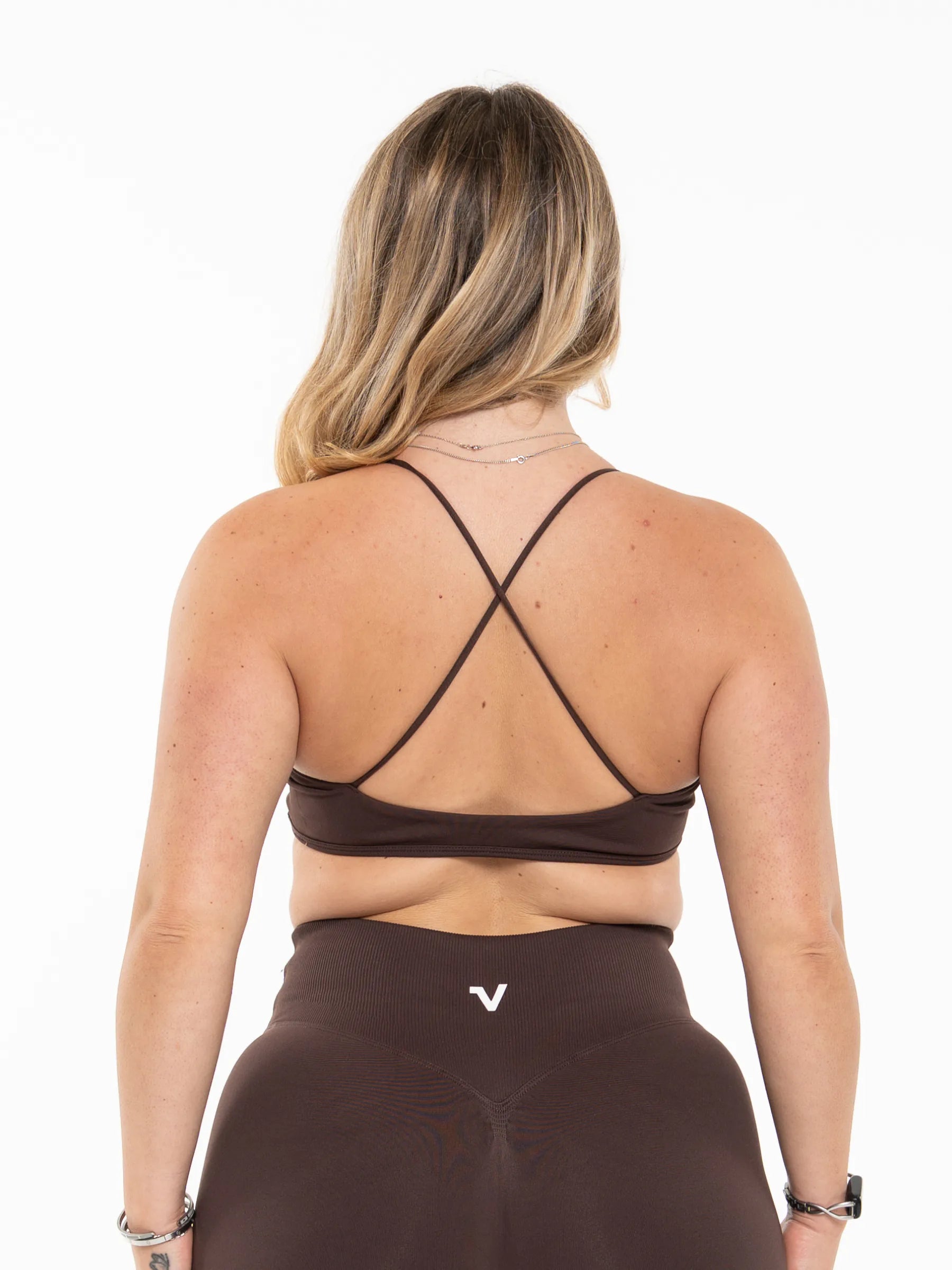 Veritone Ease Bra, Mocha Brown – A woman viewed from the back wearing a dark brown sports bra with thin crisscross straps and matching high-waisted leggings with a small white logo, against a plain white background.