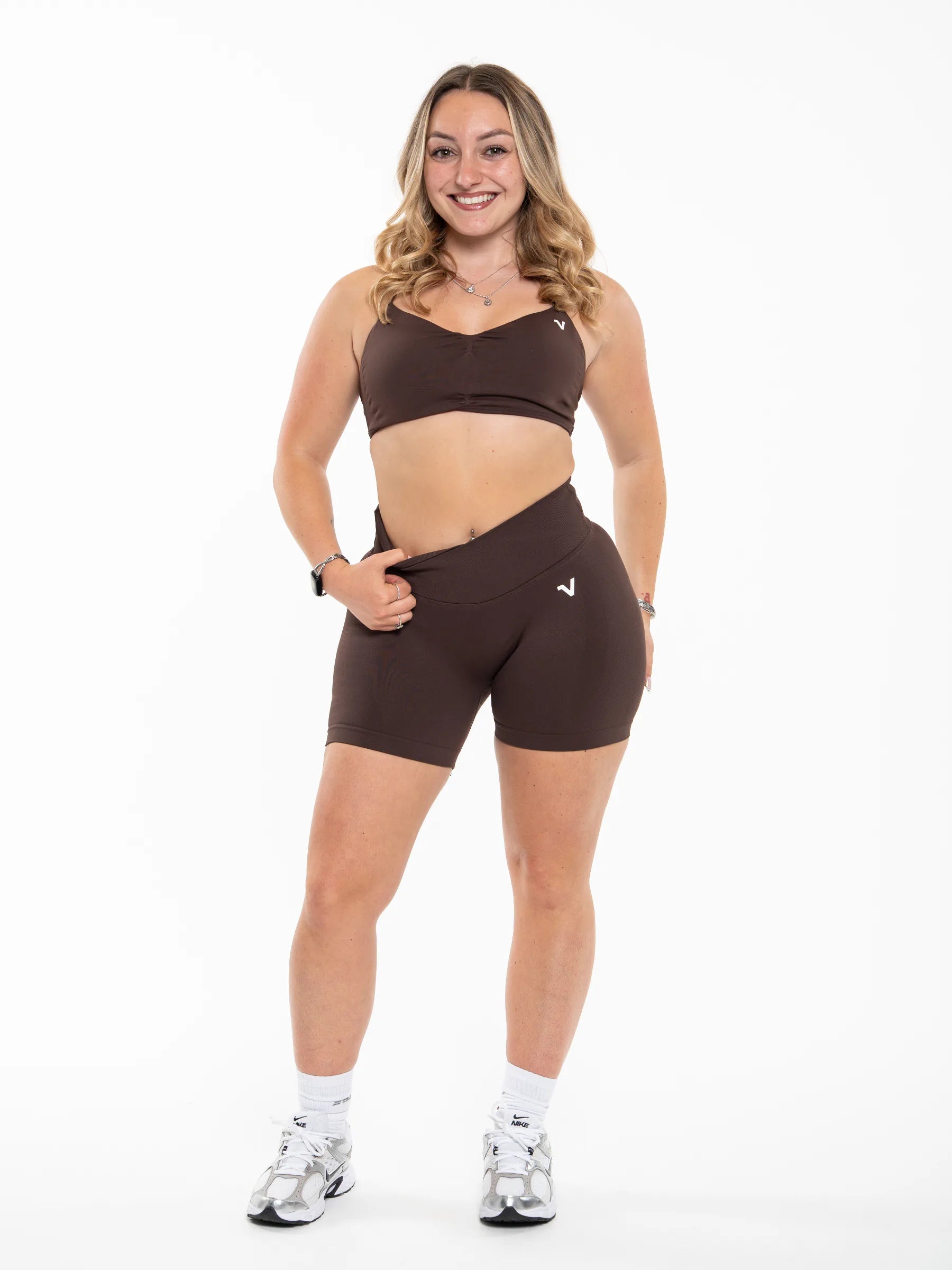 Veritone Ease Bra, Mocha Brown – A woman modeling a matching dark brown activewear set consisting of a sports bra and high-waisted shorts, paired with white socks and athletic shoes, against a plain white background.