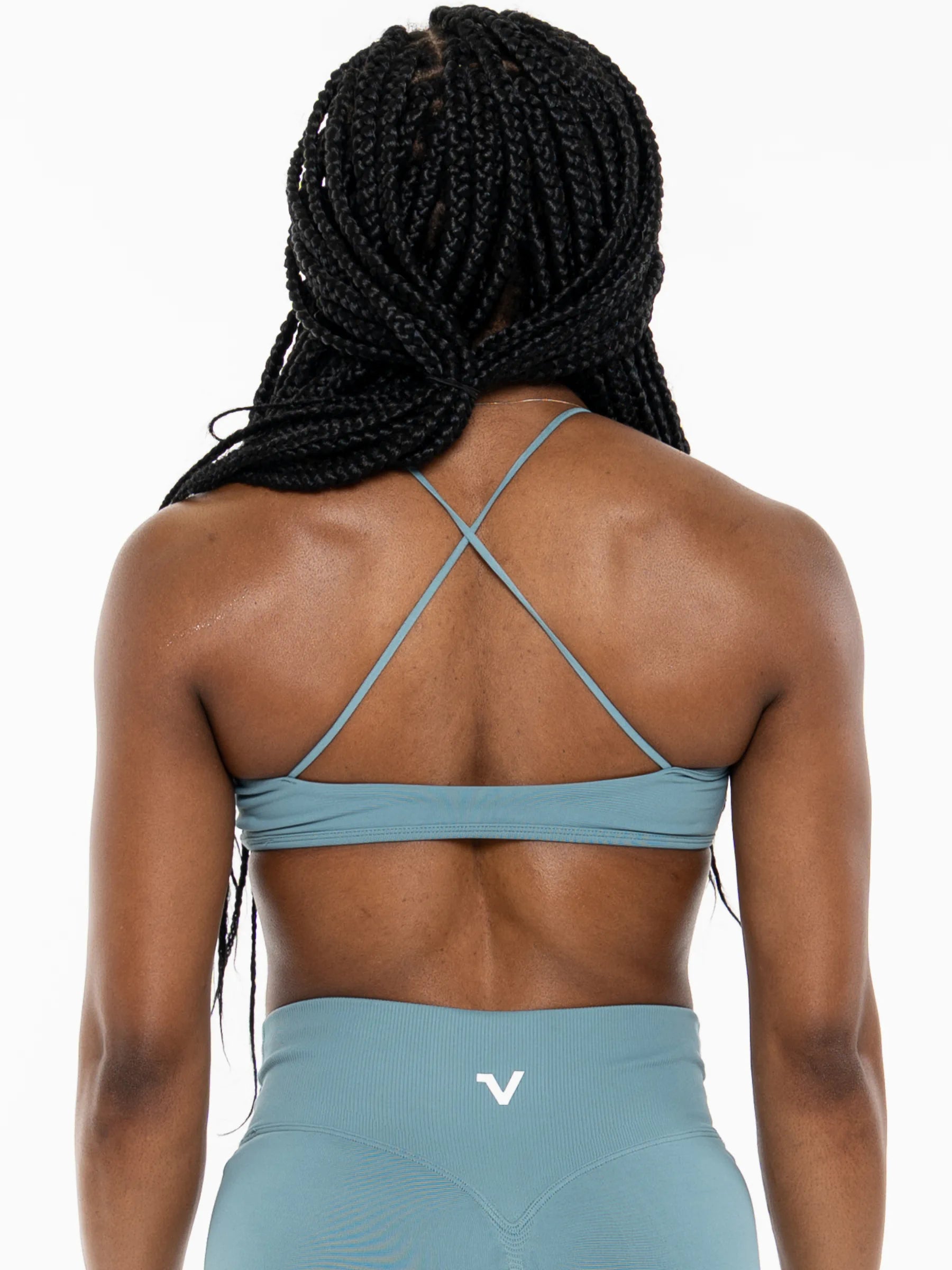 Veritone Ease Bra, Ocean Mist – Rear view of a woman with long black braids wearing a dusty blue athletic sports bra with crisscross back straps and matching high-waisted leggings with a white logo.