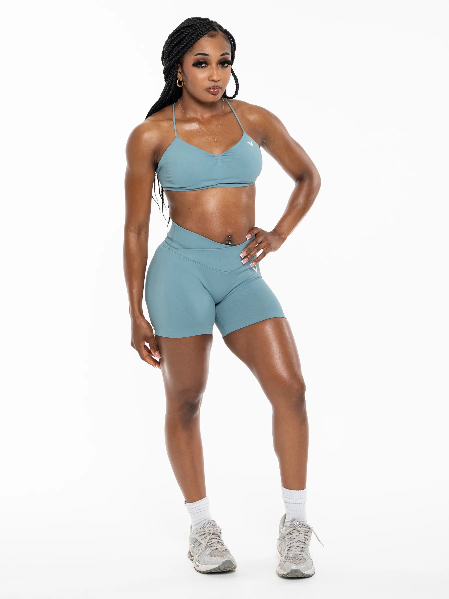 Veritone Ease Bra, Ocean Mist – A woman modeling a matching athletic set consisting of a dusty blue sports bra and high-waisted shorts, paired with white socks and gray sneakers, against a plain white studio background.
