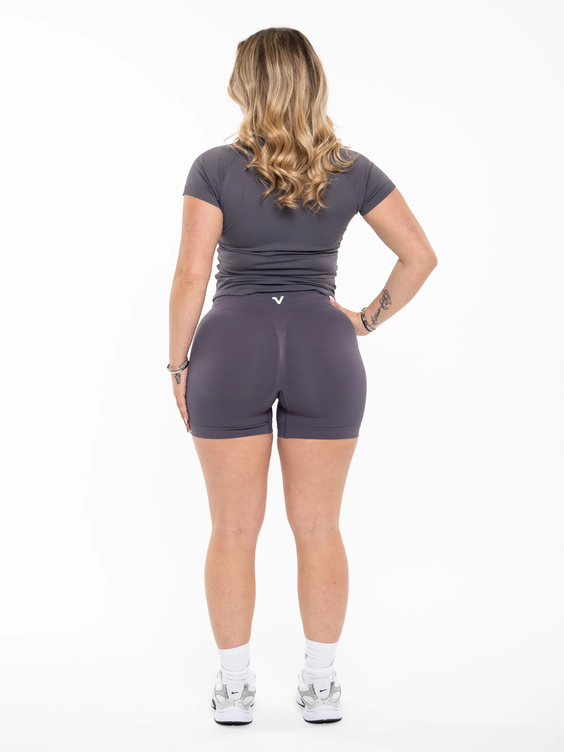 Veritone Ease Shorts, Graphite Grey – A woman is shown from the back wearing a dark gray short-sleeve athletic top and matching dark gray fitted shorts, paired with white socks and gray sneakers, against a plain white studio background.