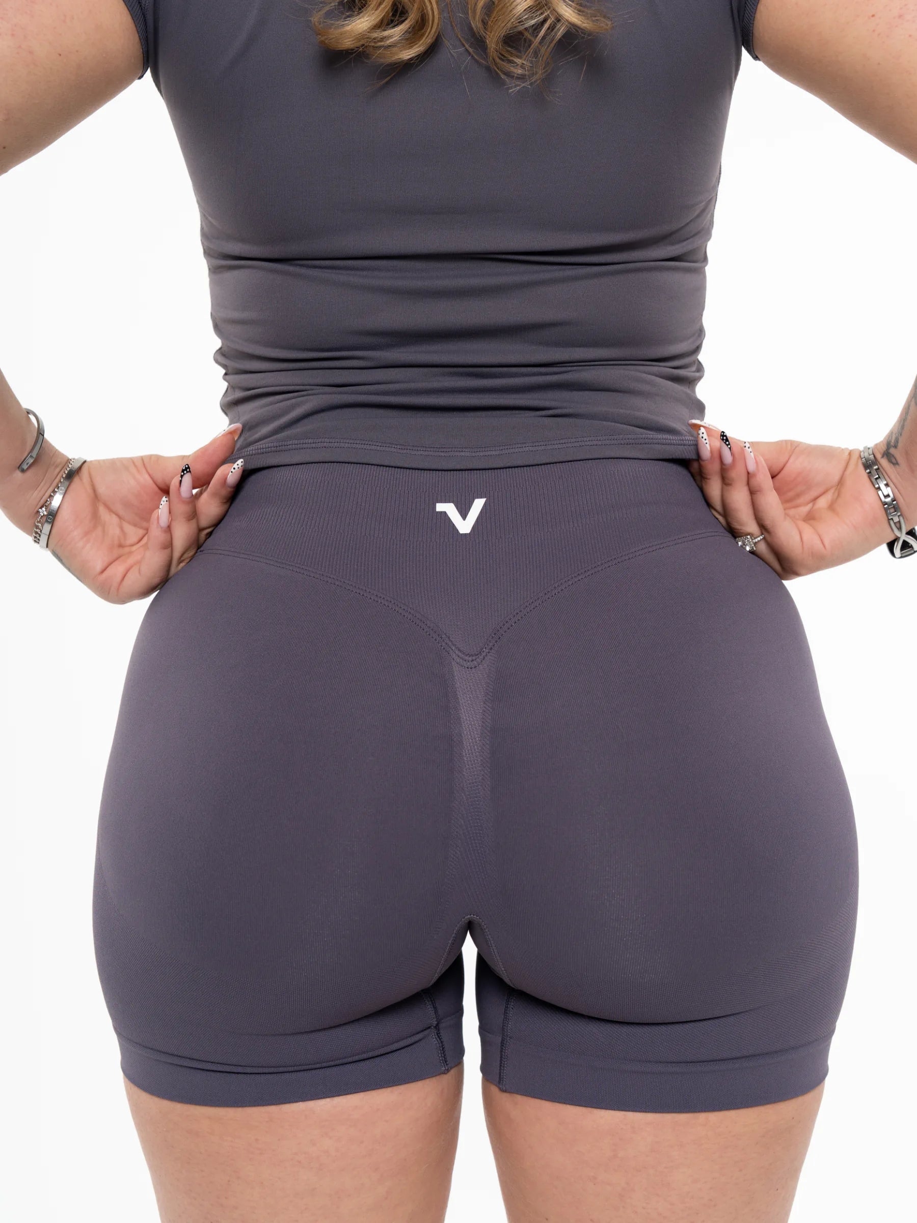 Veritone Ease Shorts, Graphite Grey – Rear view of a woman wearing dark gray athletic shorts and a matching top, with hands pulling the waistband slightly outward and a small white stylized 'V' logo centered below the waist.