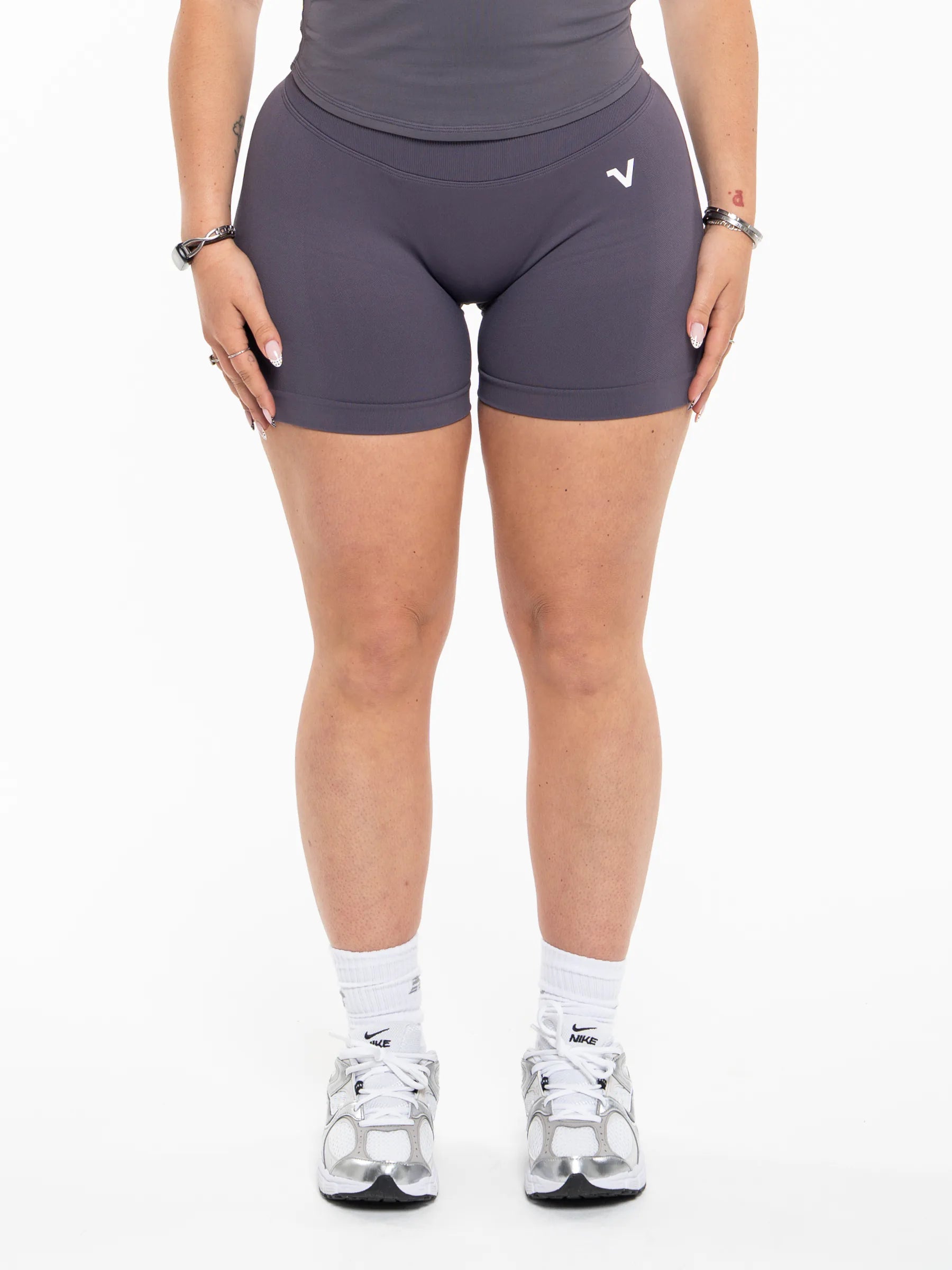Veritone Ease Shorts, Graphite Grey – A model from the waist down wearing dark gray athletic biker shorts with a small white logo on the left hip, paired with white socks and silver sneakers, against a plain white background.