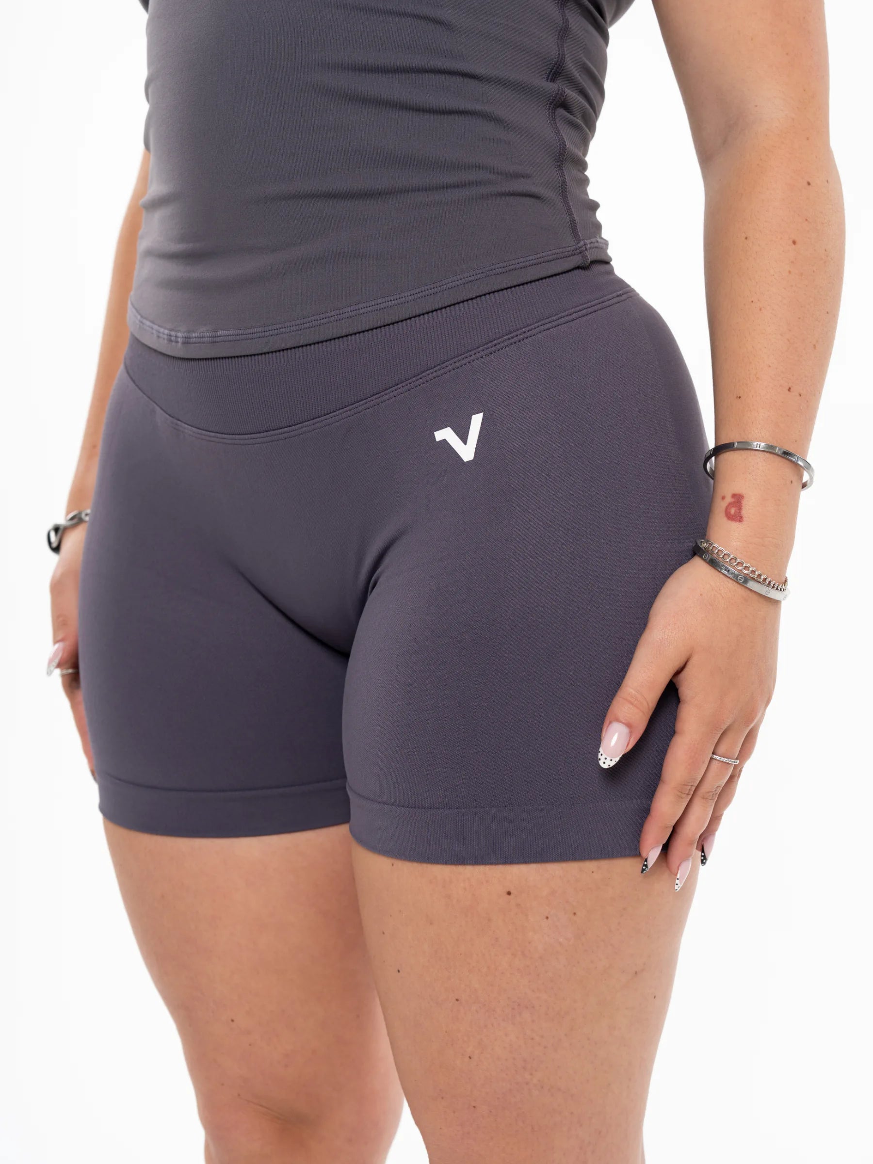 Veritone Ease Shorts, Graphite Grey – A close-up of a model wearing dark gray athletic compression shorts with a small white stylized 'V' logo on the left hip, paired with a matching dark gray top against a plain white background.