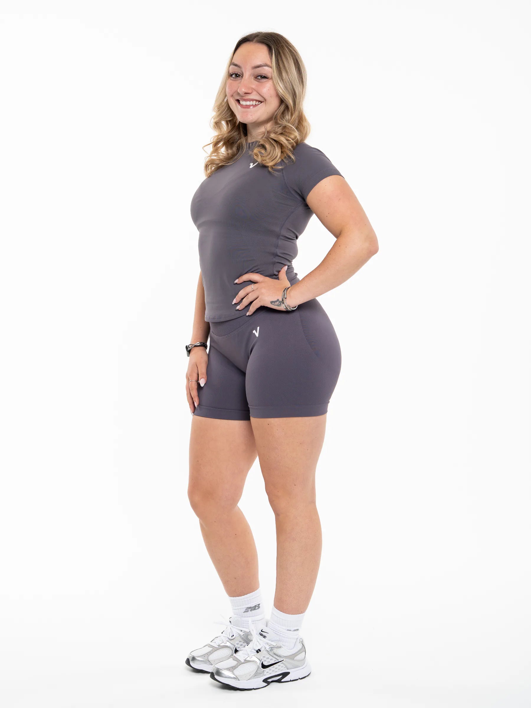 Veritone Ease Shorts, Graphite Grey – A woman modeling a matching dark gray athletic set consisting of a short-sleeve fitted top and high-waisted shorts, paired with white socks and silver sneakers, against a plain white background.