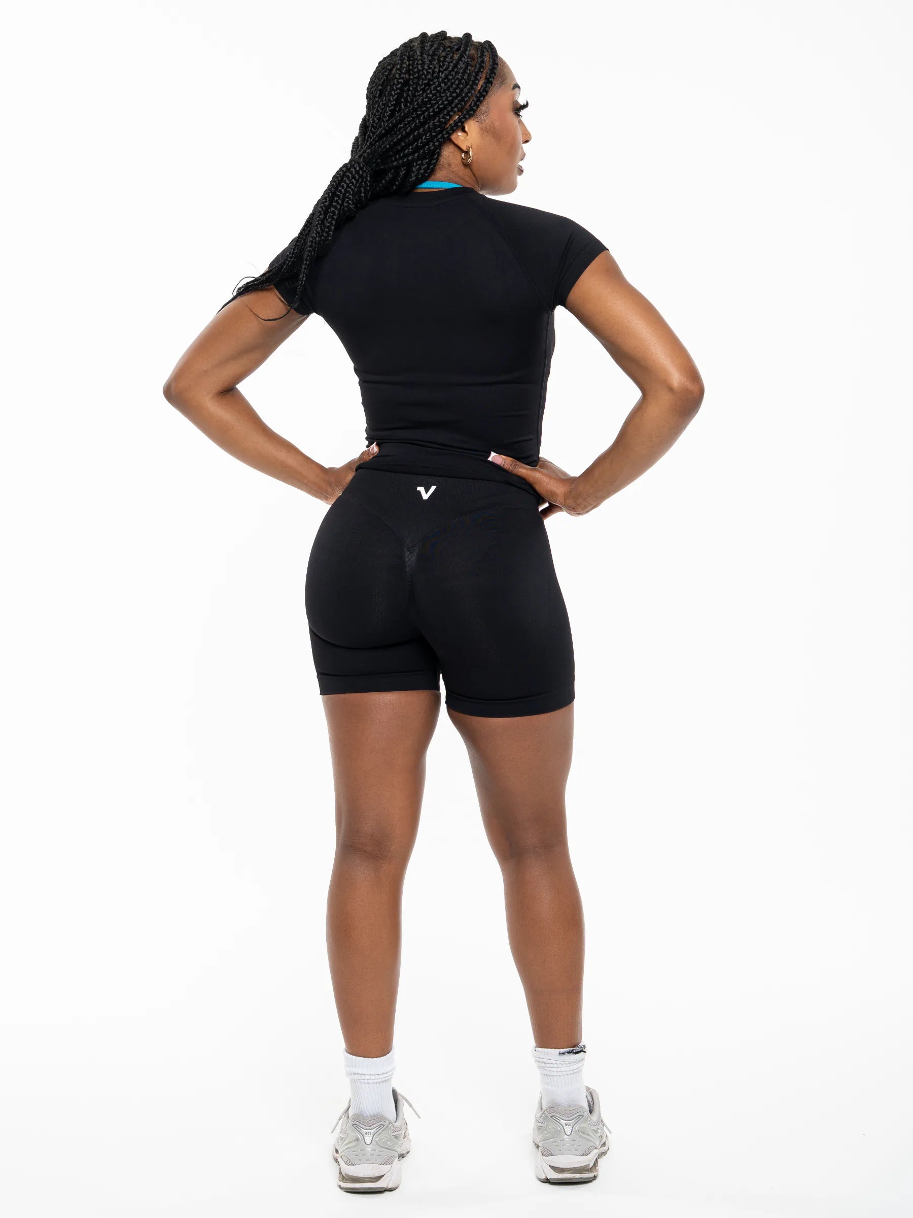 Veritone Ease Shorts, Midnight Black – A woman shown from the back wearing a fitted black athletic outfit consisting of a short-sleeve top and high-waisted biker shorts with a small white logo near the back waistband. She is standing against a plain white studio background with hands on hips.