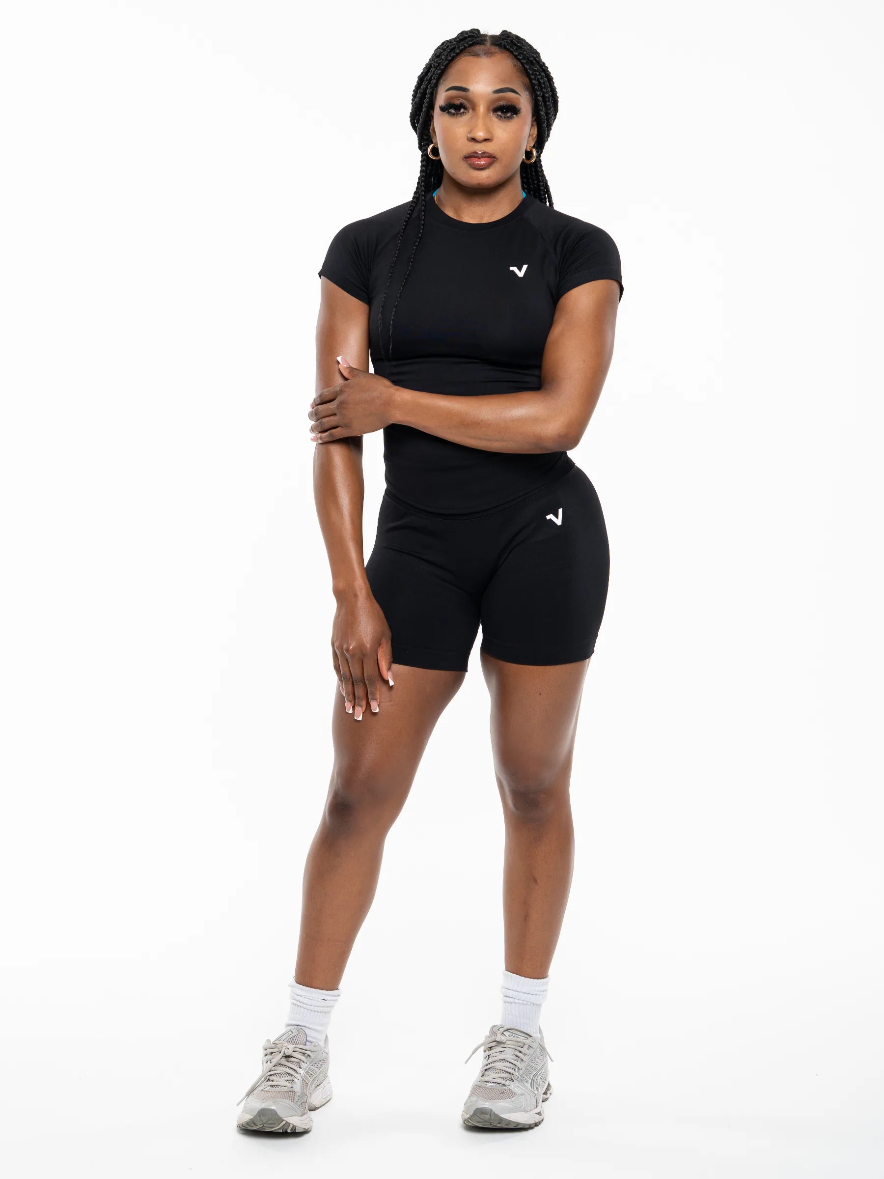 Veritone Ease Shorts, Midnight Black – A woman modeling a matching black athletic set consisting of a short-sleeve crewneck top and high-waisted shorts, featuring a small white wing logo on the chest and left thigh. She is wearing white socks and gray running shoes against a solid white studio background.