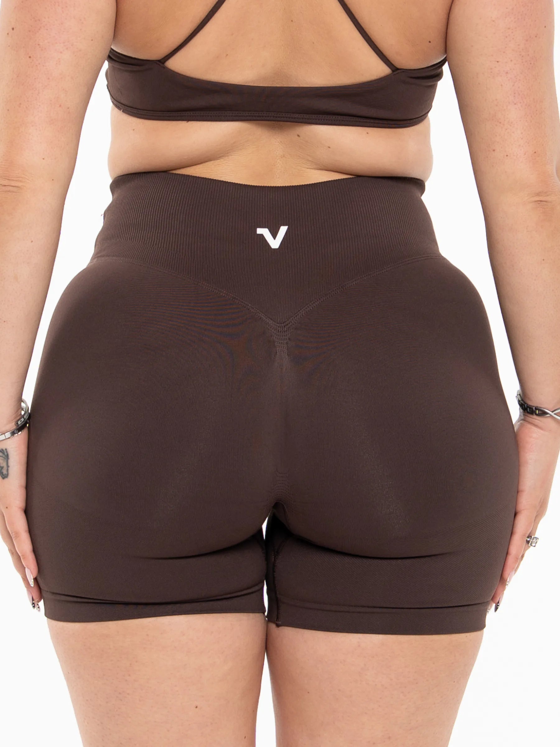 Veritone Ease Shorts, Mocha Brown – Rear view of a model wearing high-waisted brown athletic shorts with a small white logo on the center back waistband, paired with a matching brown sports bra against a plain white background.