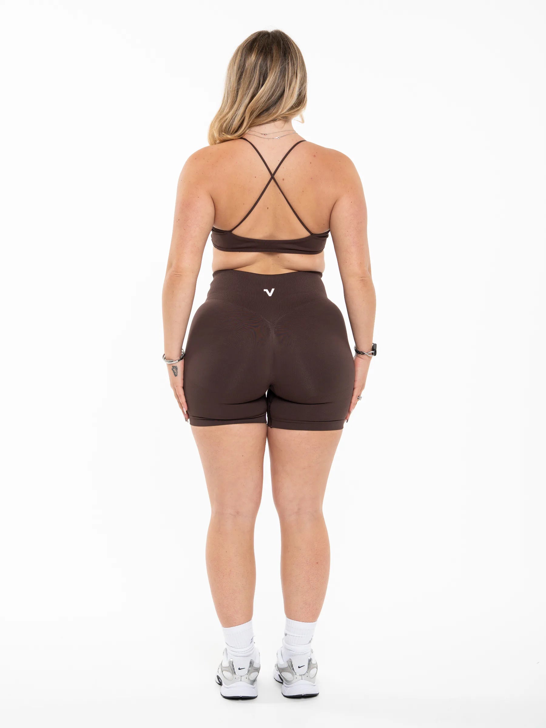 Veritone Ease Shorts, Mocha Brown – A woman viewed from the back wearing a dark brown athletic sports bra with crisscross straps and matching high-waisted dark brown shorts, paired with white socks and gray sneakers, against a plain white studio background.