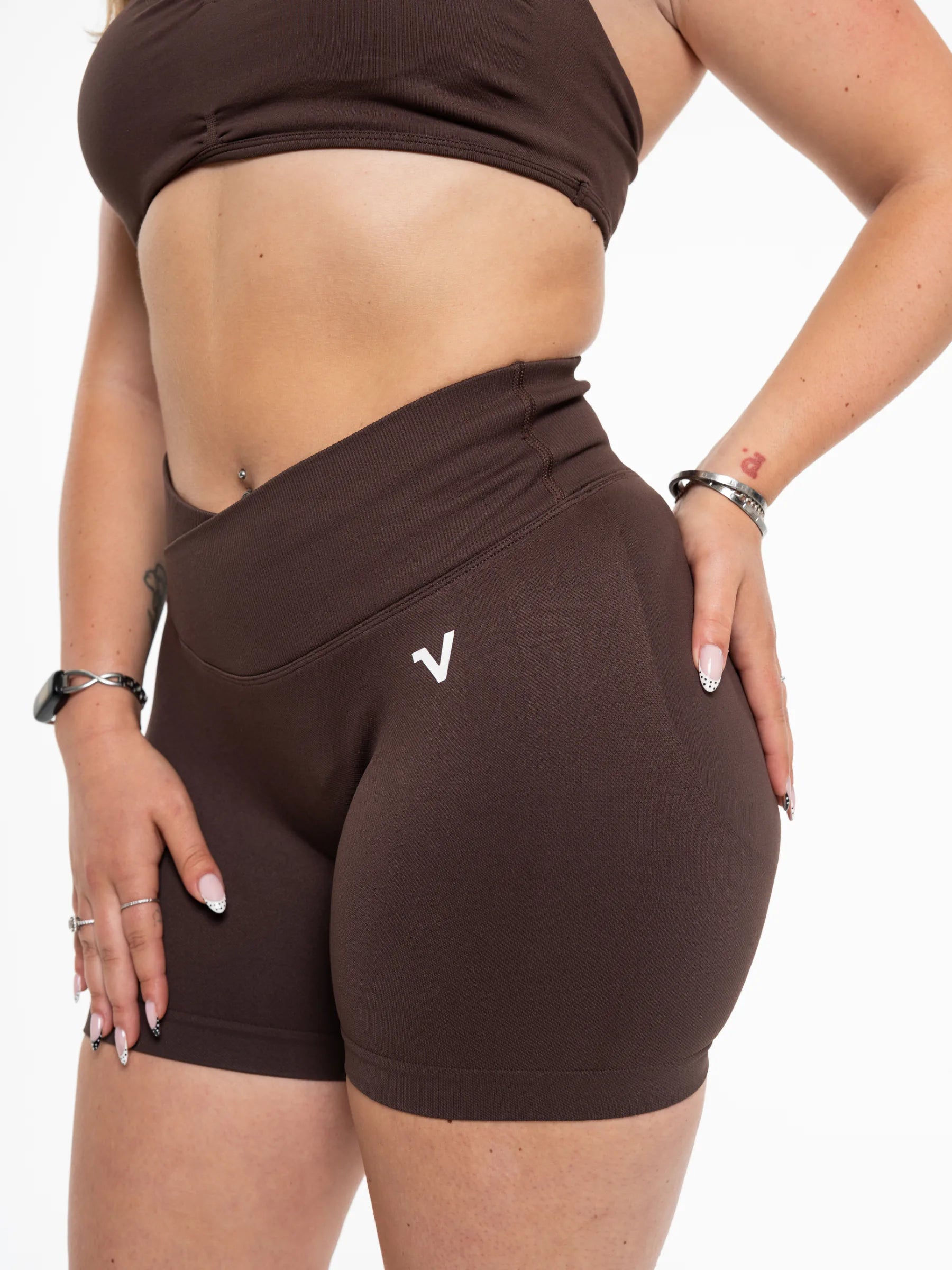 Veritone Ease Shorts, Mocha Brown – A close-up of a model wearing dark brown athletic shorts with a small white stylized 'V' logo on the left hip, paired with a matching dark brown sports bra against a plain white background.