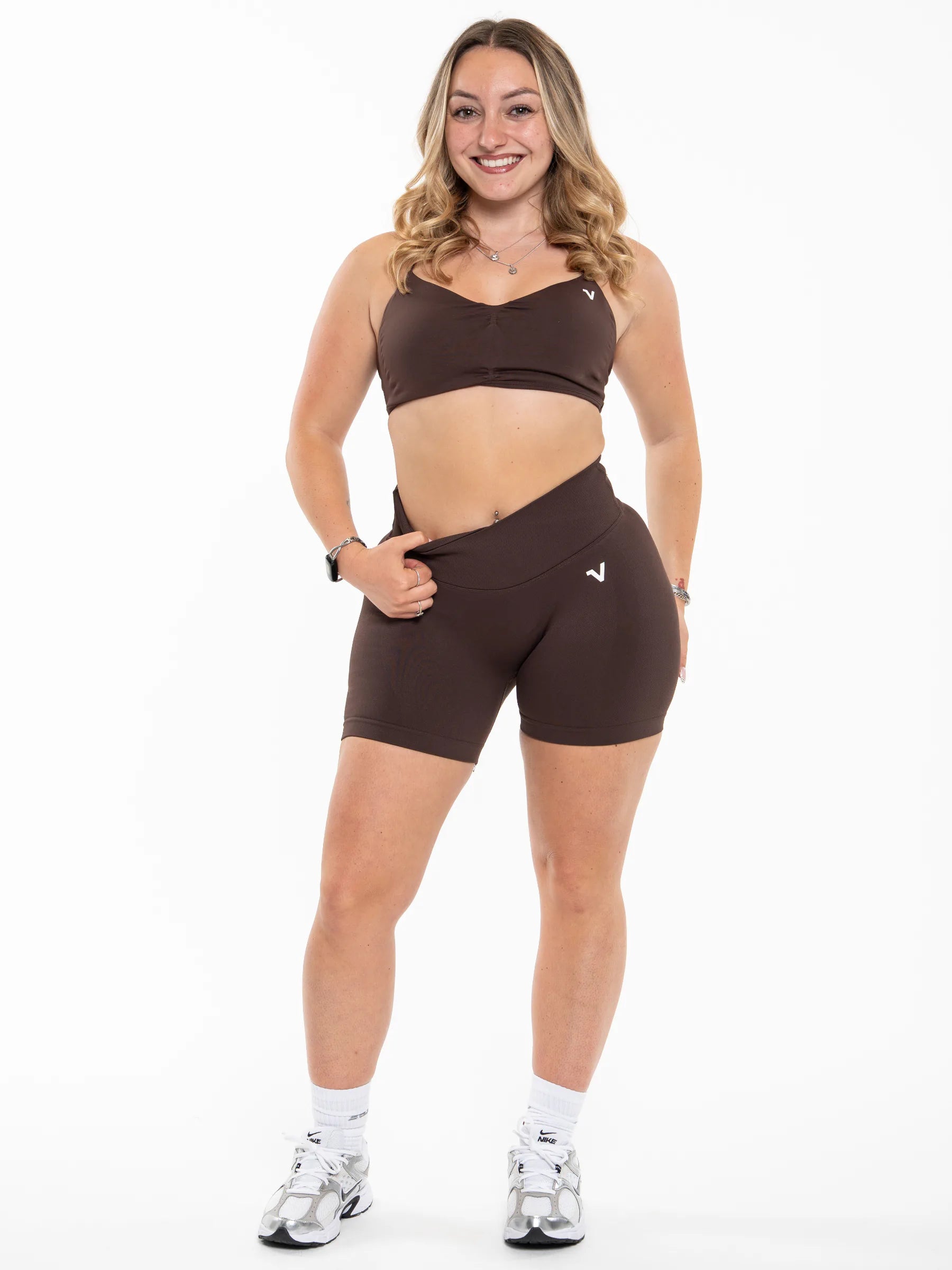 Veritone Ease Shorts, Mocha Brown – A woman modeling a matching dark brown activewear set consisting of a sports bra and high-waisted shorts, paired with white socks and athletic sneakers, against a solid white background.