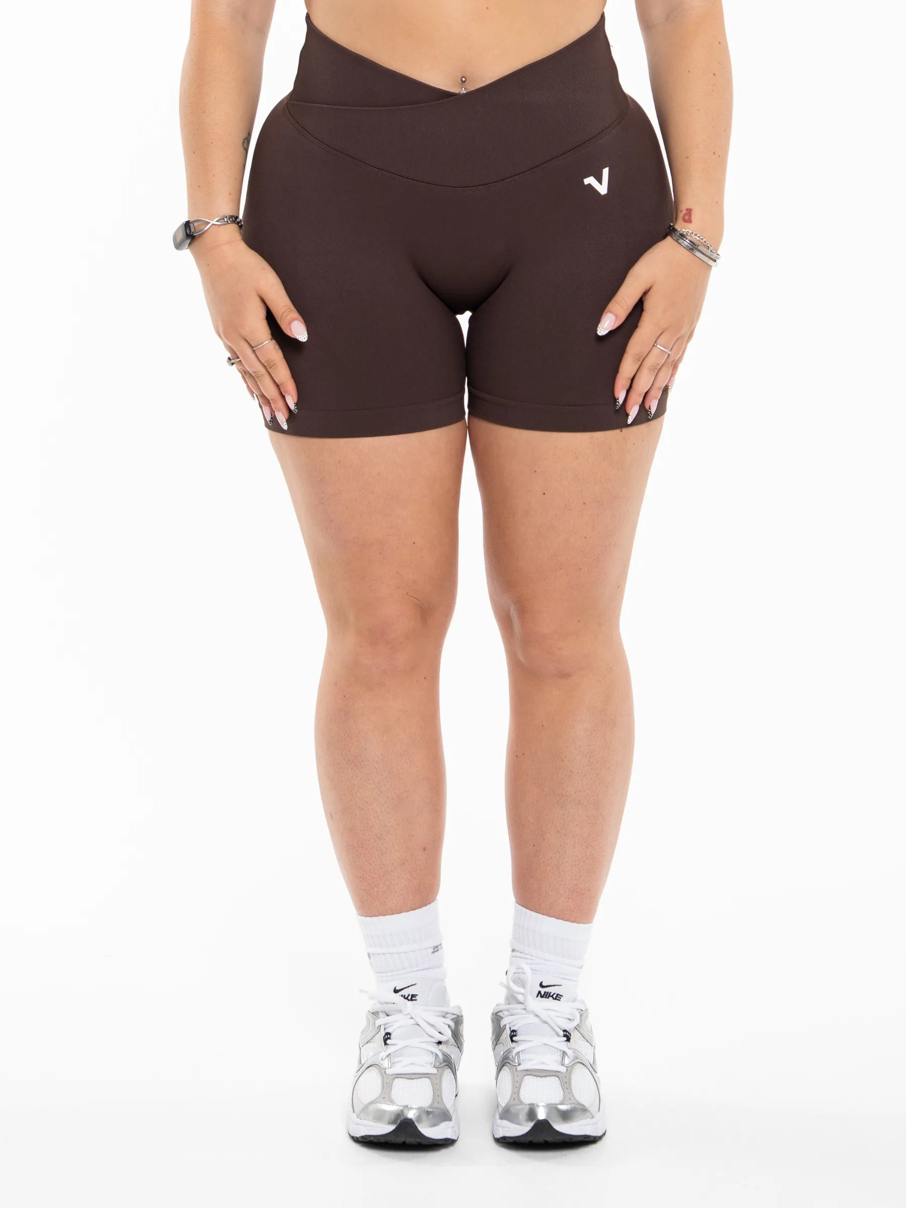 Veritone Ease Shorts, Mocha Brown – A model from the waist down wearing dark brown high-waisted biker shorts with a small white logo on the left hip, paired with white socks and silver sneakers, against a plain white background.