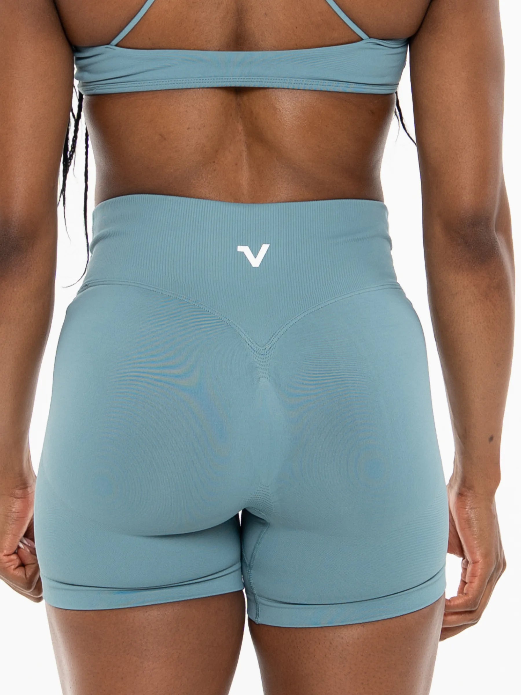 Veritone Ease Shorts, Ocean Mist – Rear view of a model wearing high-waisted athletic shorts in a dusty blue color, featuring a small white stylized 'V' logo on the center back waistband.