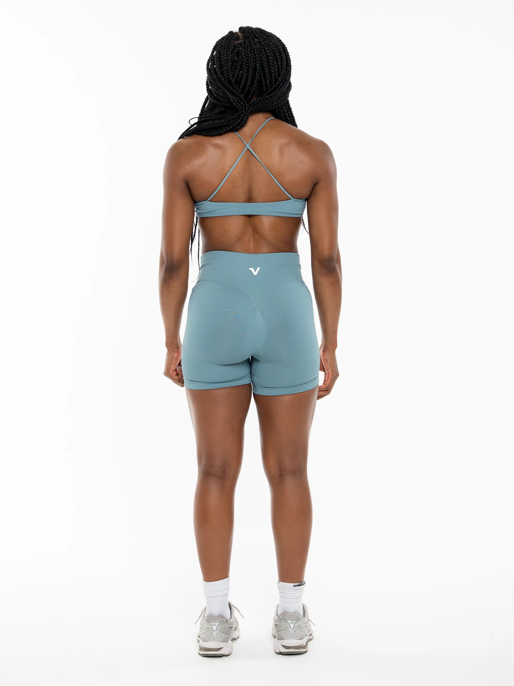 Veritone Ease Shorts, Ocean Mist – A woman viewed from behind wearing a matching dusty blue-green activewear set consisting of a sports bra with crisscross back straps and high-waisted shorts, paired with white socks and gray sneakers against a plain white background.