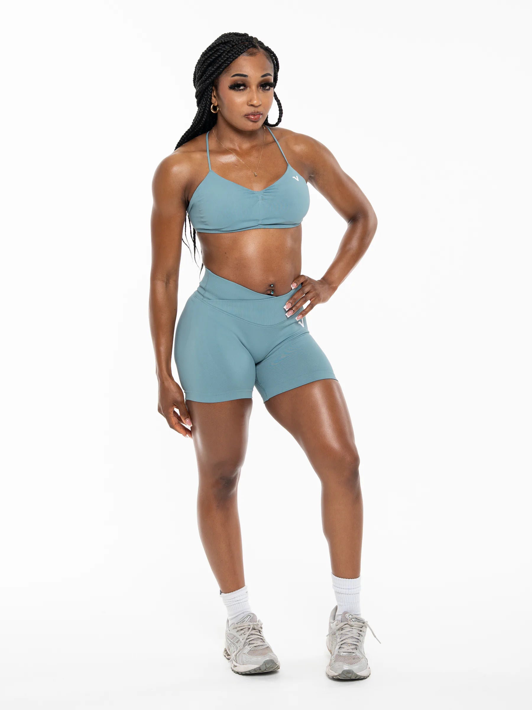 Veritone Ease Shorts, Ocean Mist – A woman modeling a matching athletic set consisting of a dusty blue sports bra and high-waisted shorts, paired with white socks and gray sneakers, against a plain white studio background.