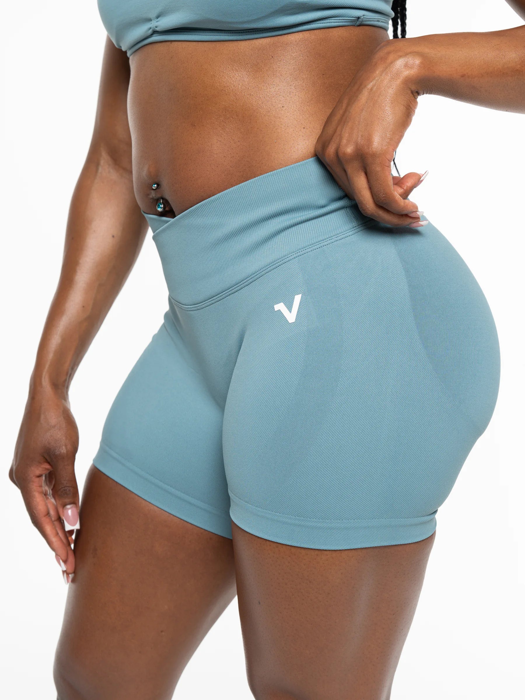 Veritone Ease Shorts, Ocean Mist – A close-up of a model wearing high-waisted, cheeky athletic shorts in a dusty blue color, featuring a white stylized 'V' logo on the left hip.