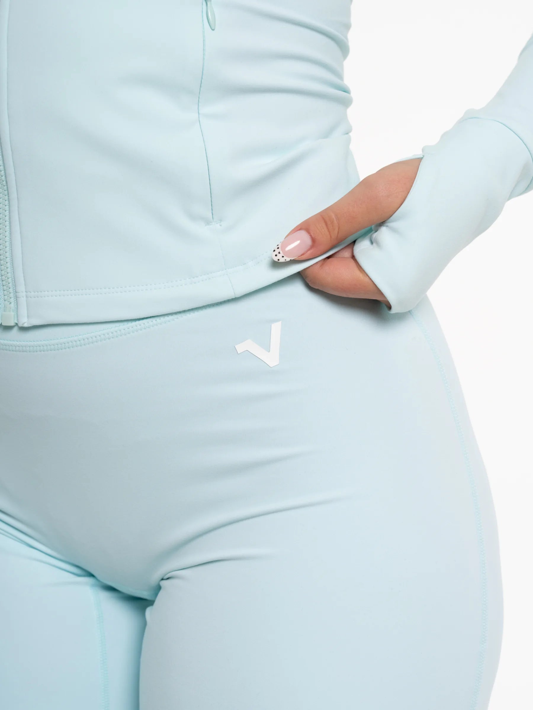 Veritone Élan Flared Pants, Ocean Mist – Close-up of a person wearing a light pastel blue athletic zip jacket and matching leggings, with a white stylized V logo 