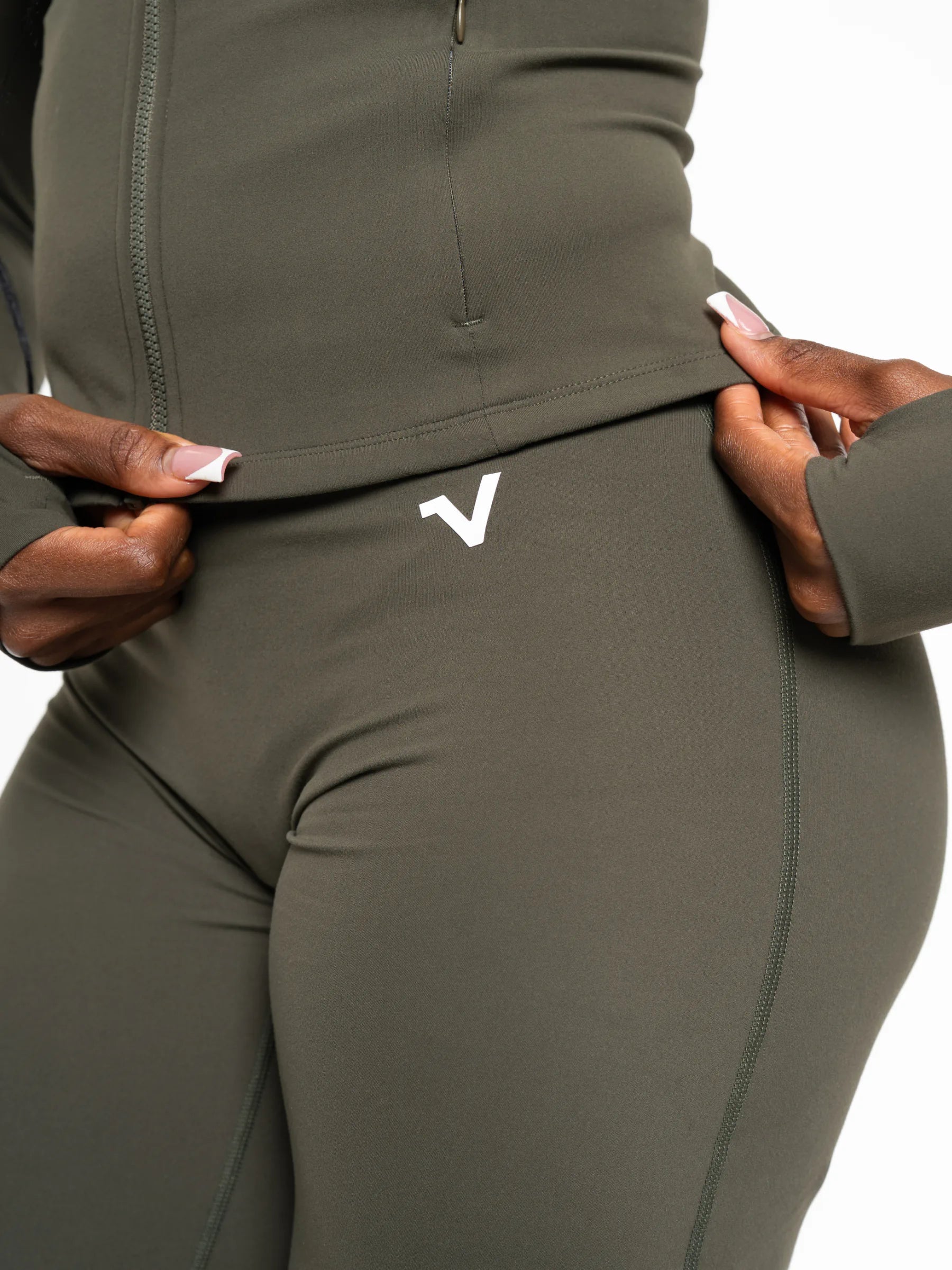 Veritone Élan Flared Pants, Olive Stone – Close-up of a person wearing olive green athletic leggings and a matching zip-up top, pulling the waistband slightly outward with both hands. A small white stylized 'V' logo is visible on the hip.