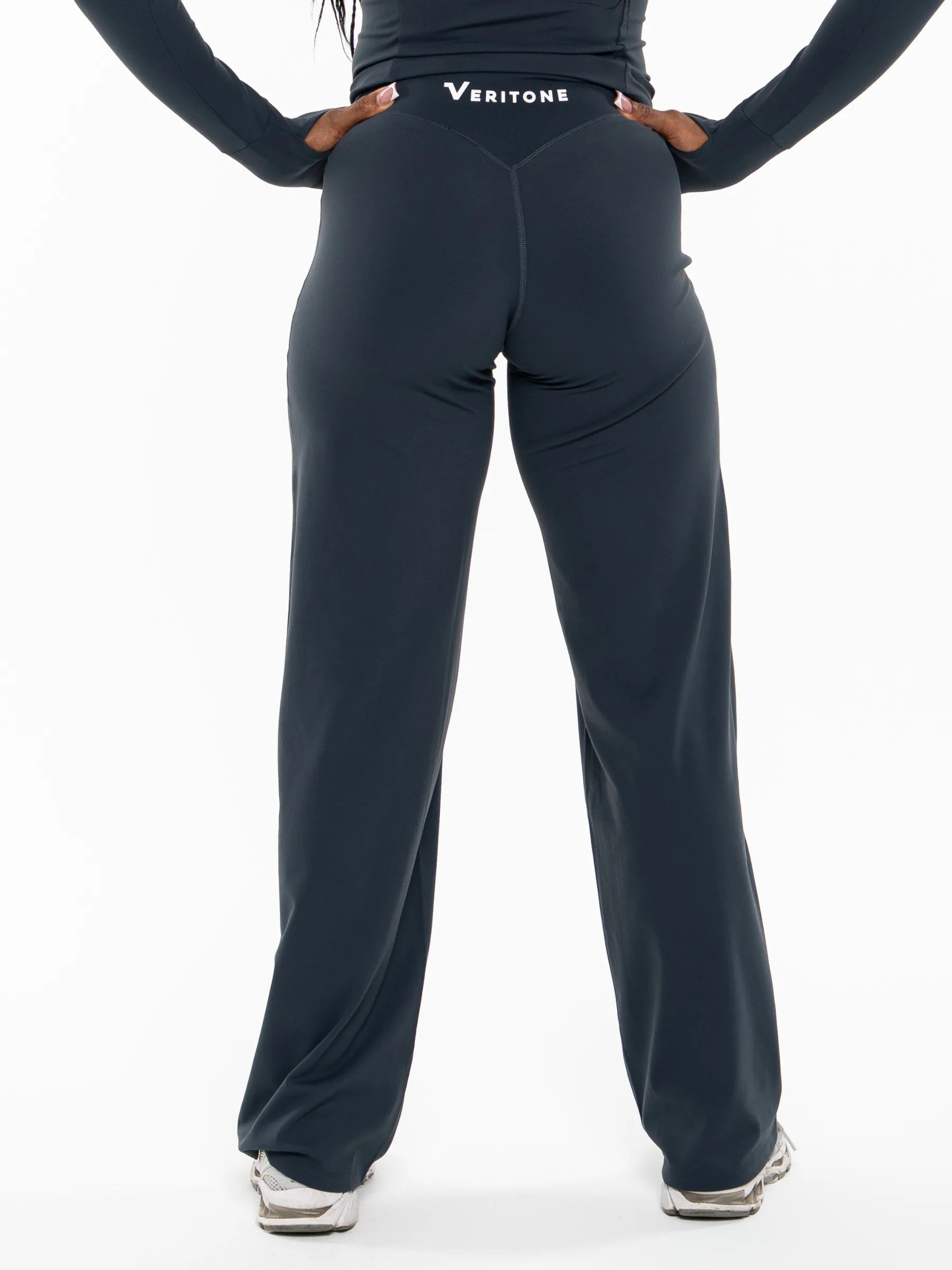Veritone Élan Flared Pants, Storm Blue – Rear view of a person wearing dark navy wide-leg athletic pants with a visible Veritone logo at the back waistband, paired with a matching long-sleeve top and sneakers on a white studio background.