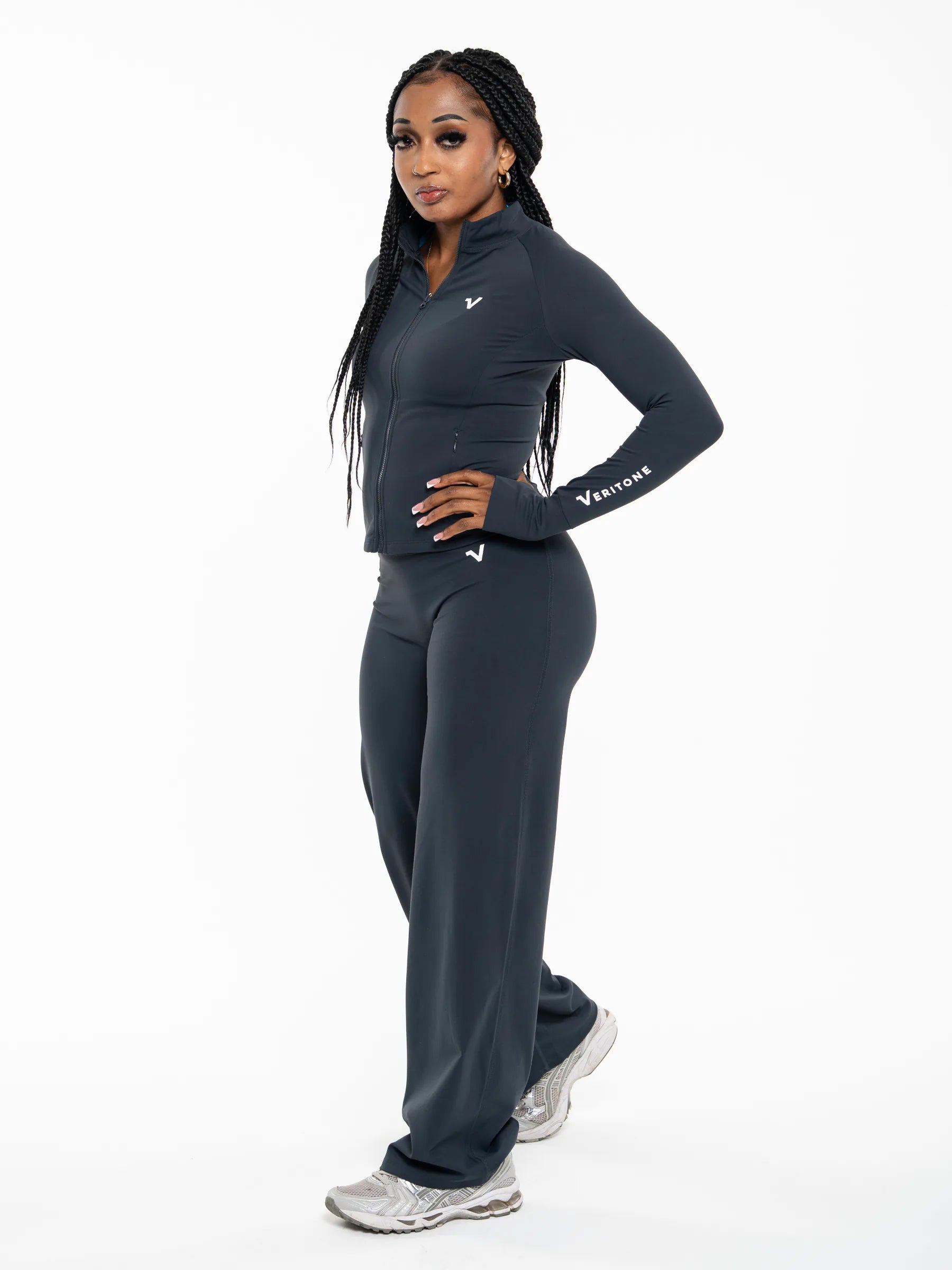 Veritone Élan Flared Pants, Storm Blue – A woman modeling a dark blue athletic tracksuit consisting of a long-sleeve zip top and matching wide-leg pants, with white sneakers, against a plain white studio background.