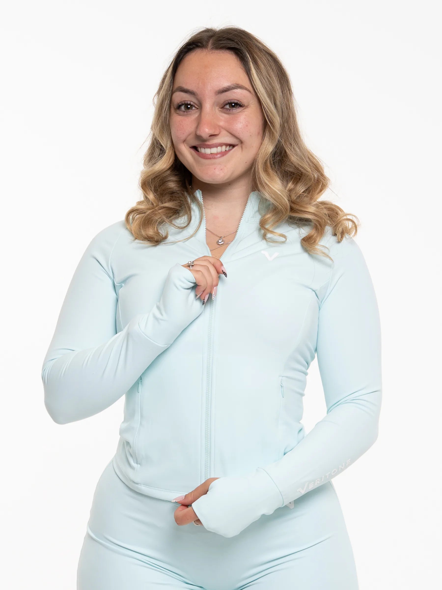 Veritone Élan Jacket, Ocean Mist – A smiling woman with blonde wavy hair wearing a light blue long-sleeve zip-up athletic jacket and matching light blue leggings, posed against a solid white studio background.