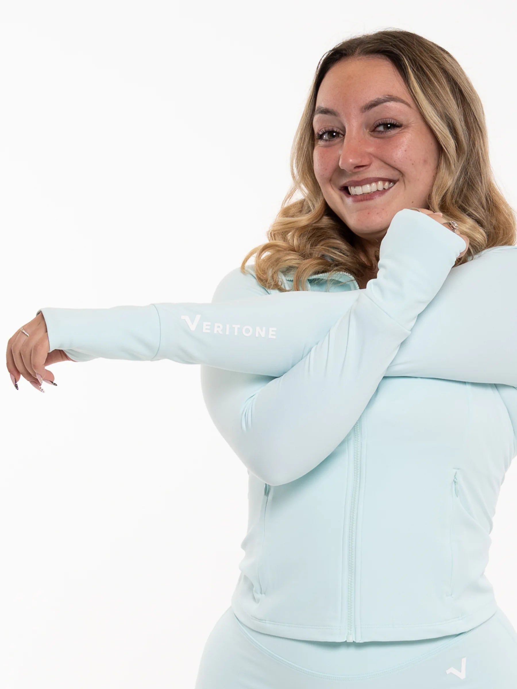 Veritone Élan Jacket, Ocean Mist – A smiling woman wearing a light pastel blue Veritone athletic zip-up jacket and matching bottoms, posing with her arms crossed against a plain white background.