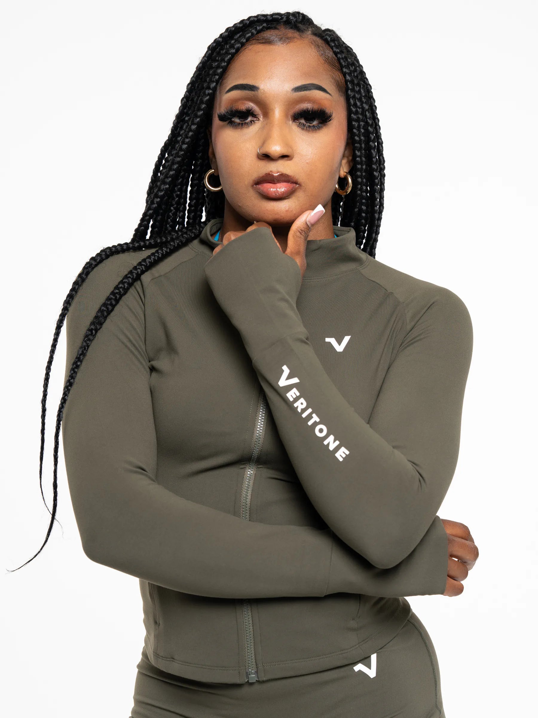 Veritone Élan Jacket, Olive Stone – A woman with long black braids wearing a fitted olive green athletic zip-up jacket and matching bottoms. The jacket has a white Veritone logo on the left chest and 'VERITONE' printed in white down the right sleeve, photographed against a plain white background.