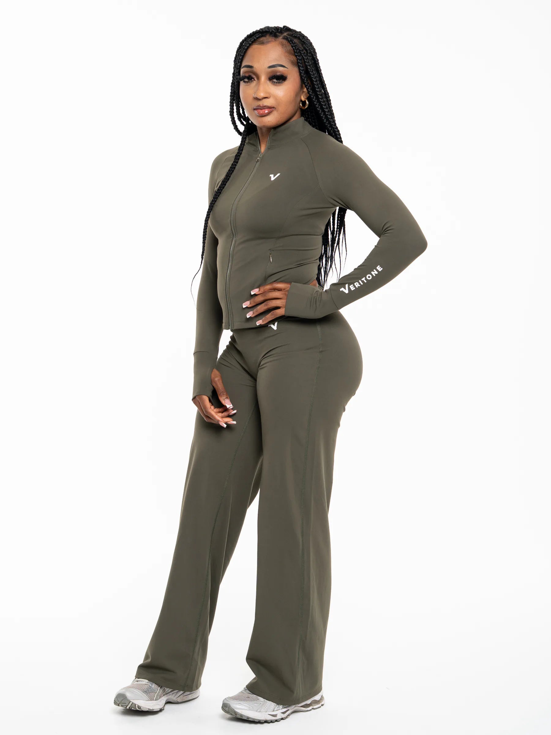 Veritone Élan Jacket, Olive Stone – A woman modeling a matching olive green athletic set consisting of a long-sleeve mock-neck zip-up top and wide-leg pants, featuring a small white logo on the chest and sleeve, set against a plain white background.