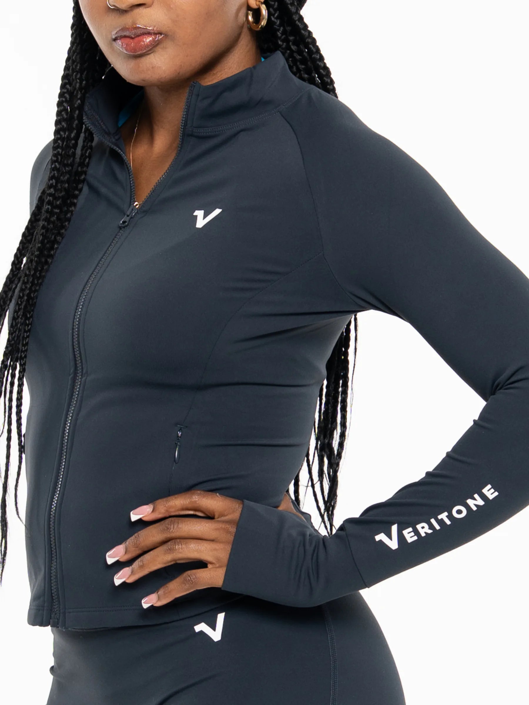 Veritone Élan Jacket, Storm Blue – A close-up of a woman wearing a dark navy athletic zip-up jacket with a white Veritone logo on the chest and sleeve, paired with matching leggings against a white background.