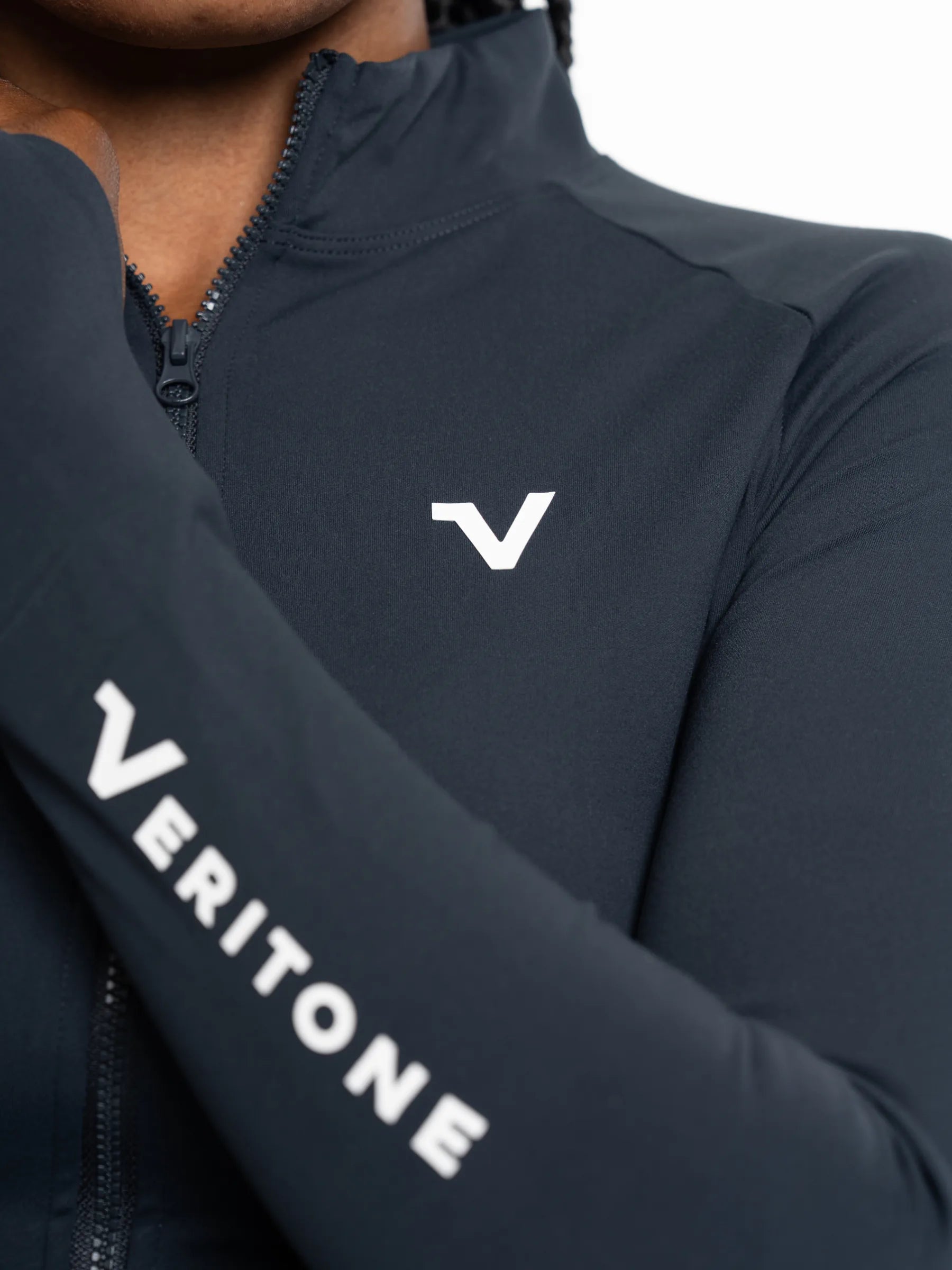 Veritone Élan Jacket, Storm Blue – Close-up of a person wearing a dark navy athletic zip-up jacket with a white Veritone logo on the chest and sleeve.