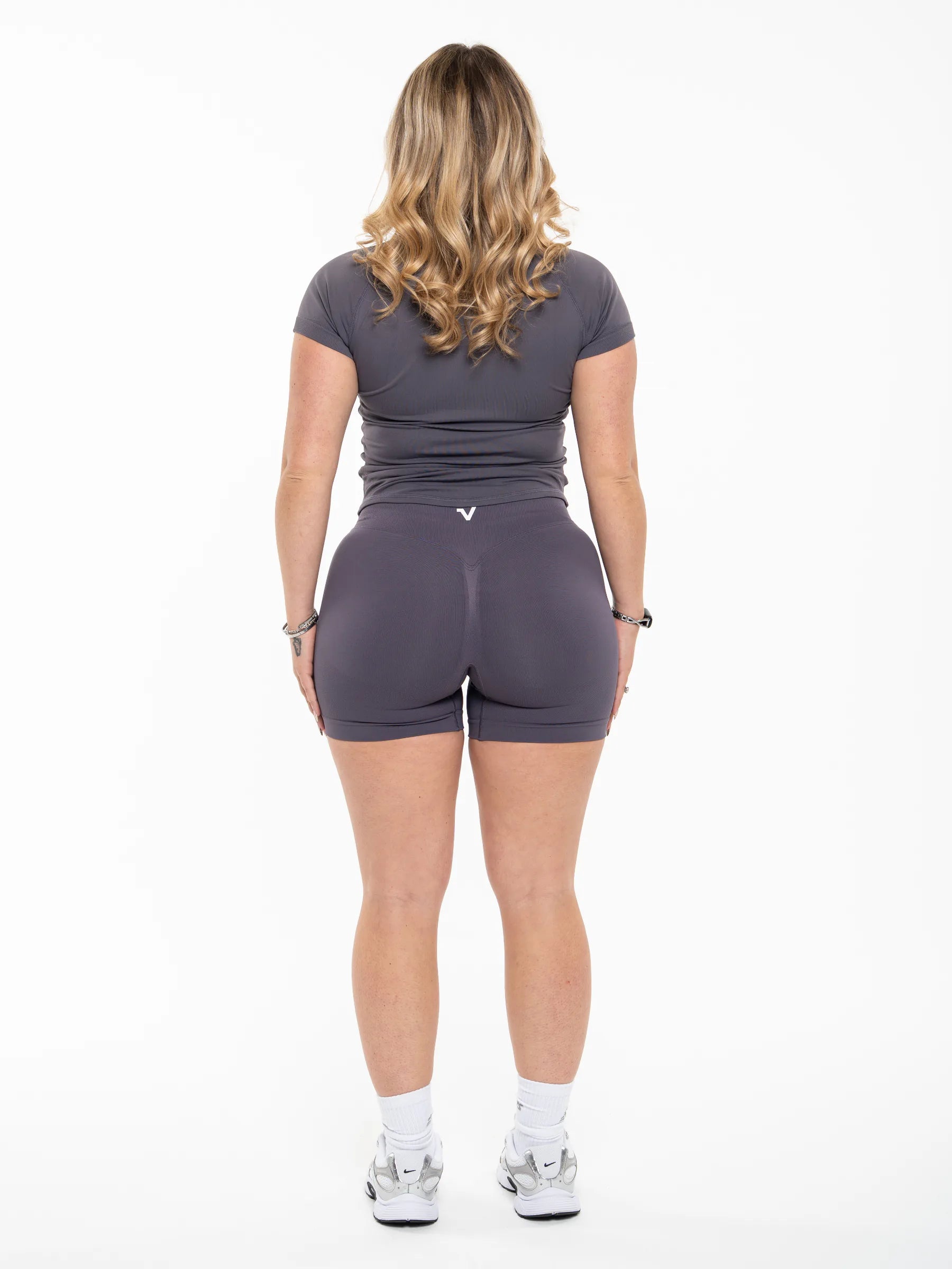 Veritone Form T-Shirt, Graphite Grey – A woman viewed from behind wearing a dark gray short-sleeve athletic top and matching high-waisted shorts, paired with white socks and gray sneakers, against a plain white studio background.