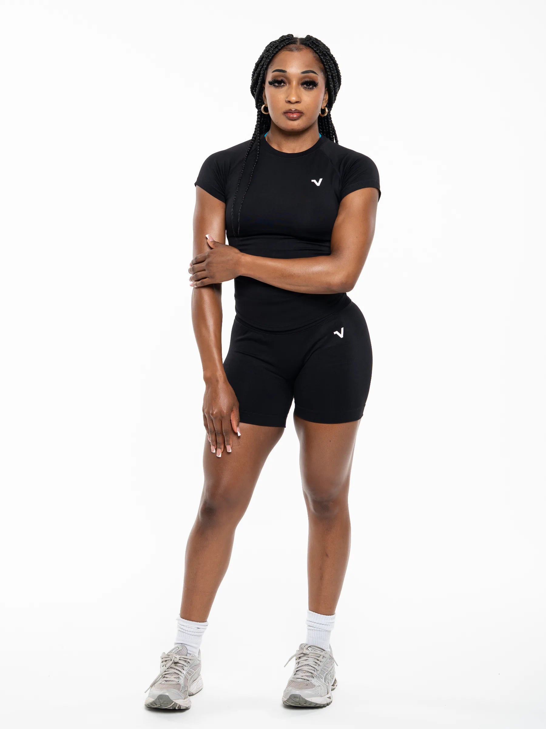 Veritone Form T-Shirt, Midnight Black – A woman modeling a black athletic set consisting of a short-sleeve crewneck shirt and matching high-waisted shorts, featuring a small white logo on the chest and left hip, paired with white socks and gray sneakers against a plain white background.