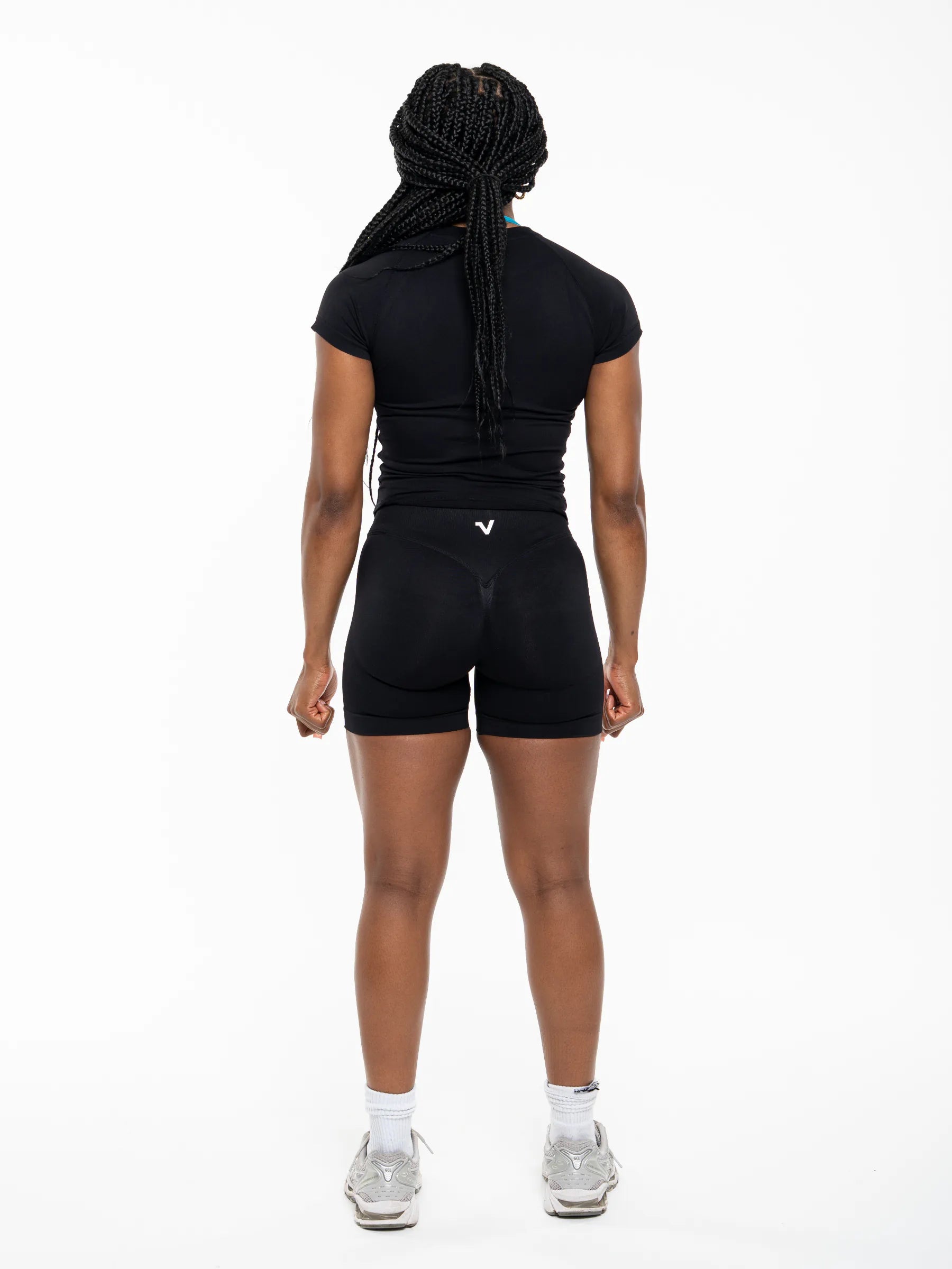 Veritone Form T-Shirt, Midnight Black – A woman viewed from the back wearing a black athletic compression outfit consisting of a short-sleeve top and fitted shorts, with white socks and gray sneakers, against a plain white studio background.