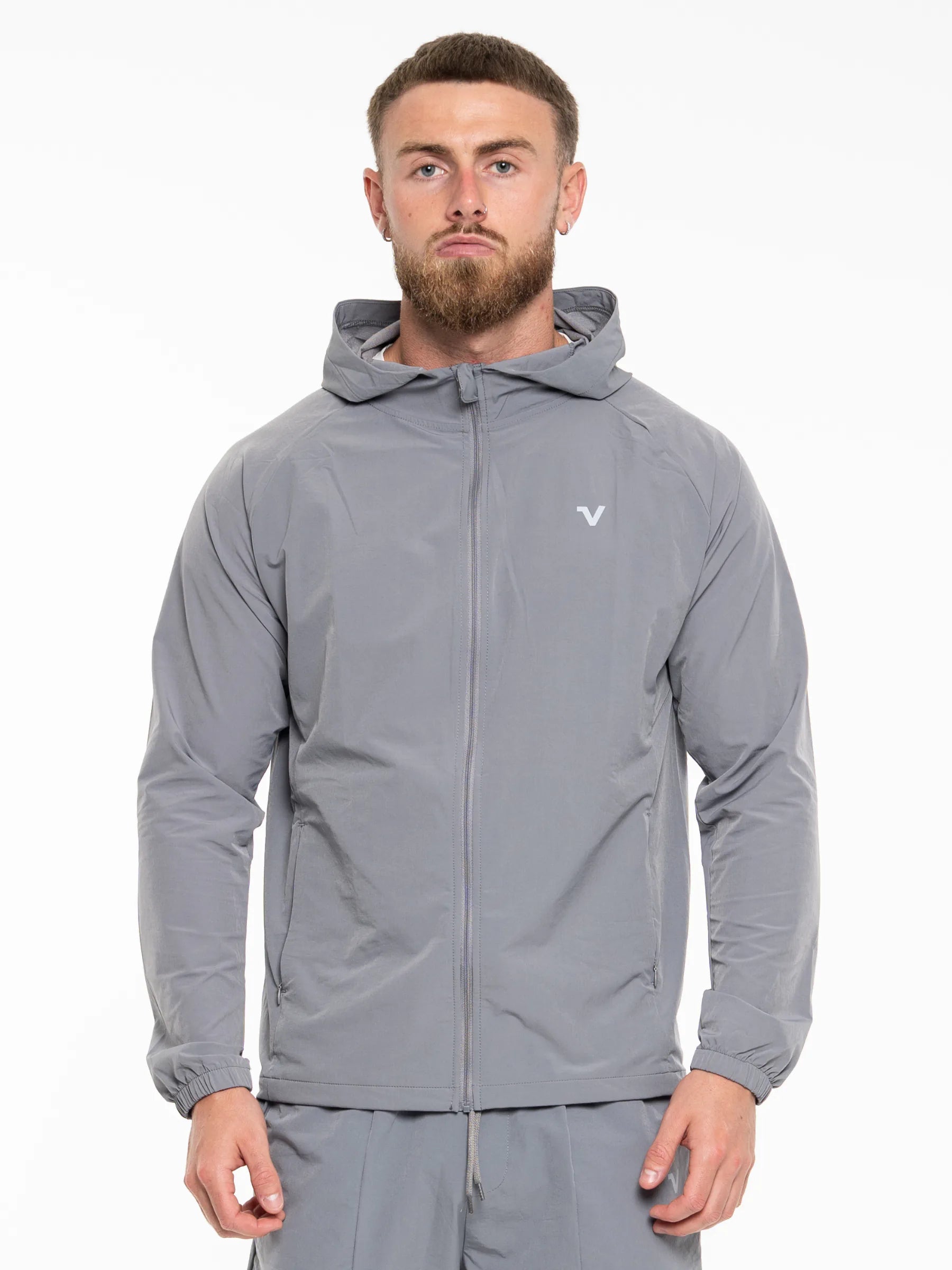 Veritone Mode Jacket, Ash Grey – A man wearing a gray hooded athletic jacket with a full front zipper and a small white logo on the left chest, paired with matching gray pants, photographed against a plain white studio background.