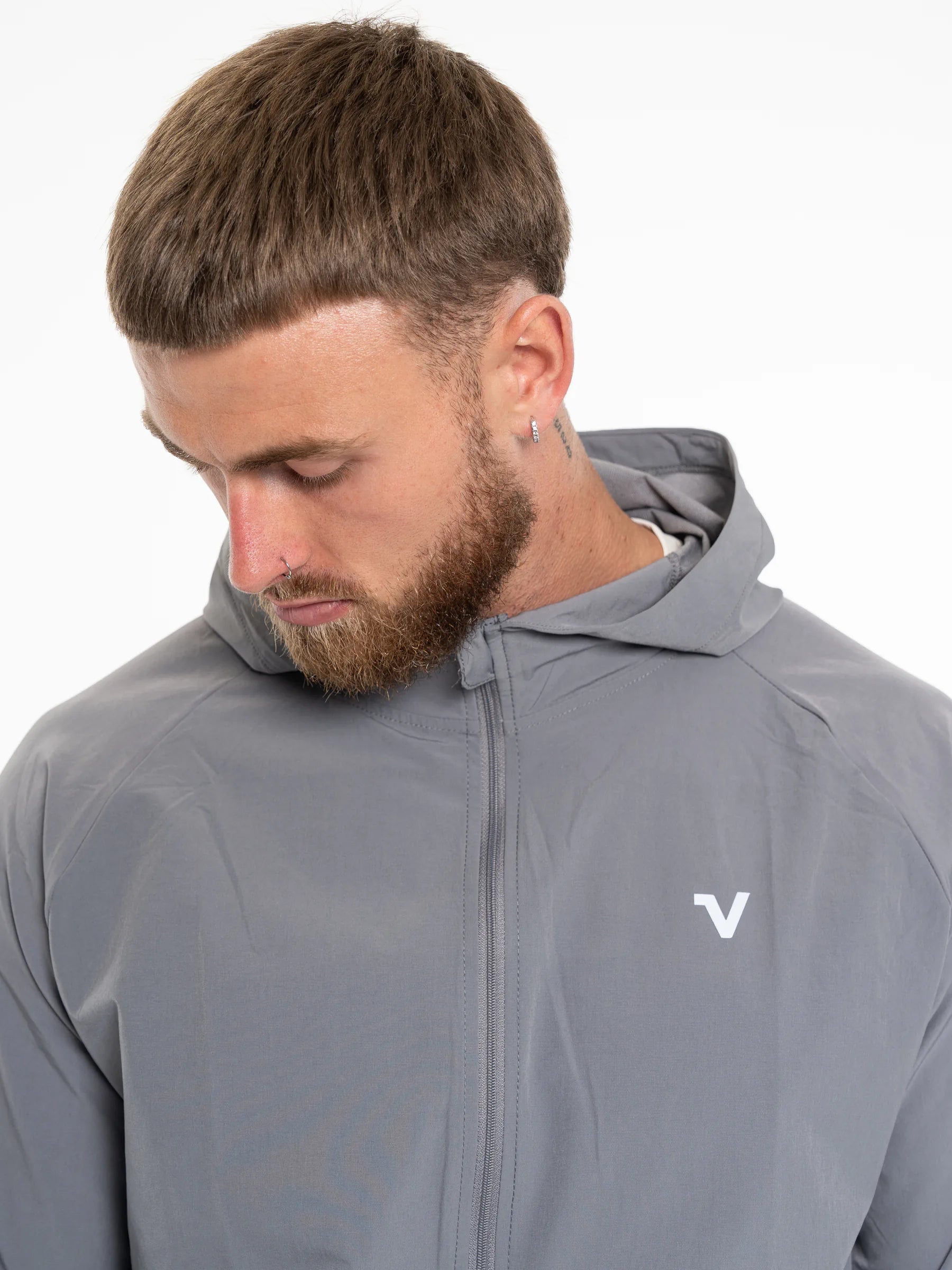 Veritone Mode Jacket, Ash Grey – A close-up shot of a man wearing a gray hooded zip-up jacket, looking down. The jacket features a small white stylized 'V' logo on the left chest, photographed against a clean white background.