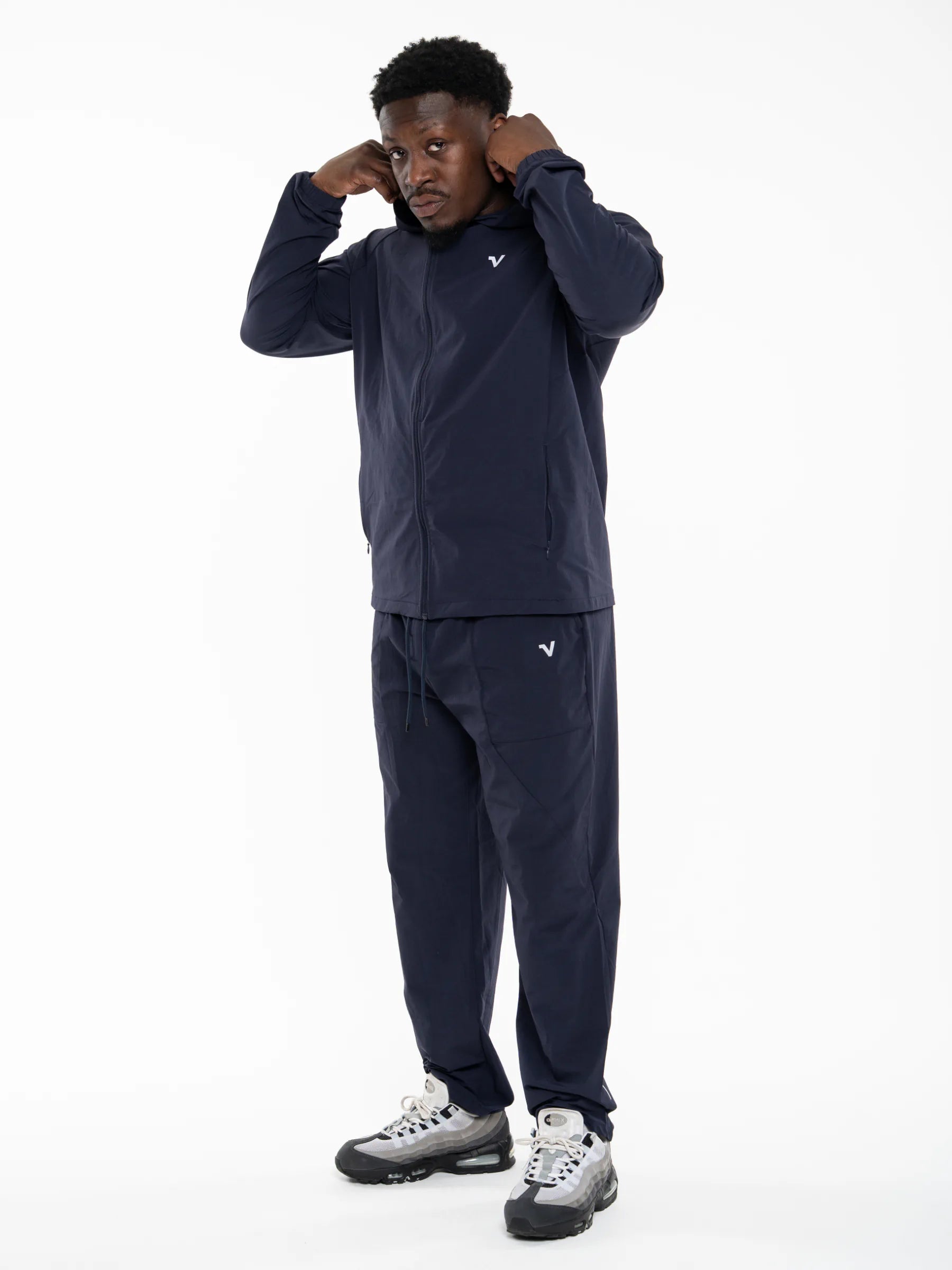 Veritone Mode Jacket, Deep Navy – A man modeling a matching navy blue athletic tracksuit, featuring a zip-up jacket and jogger pants, with a small white logo on the chest and thigh. He is standing against a solid white studio background.