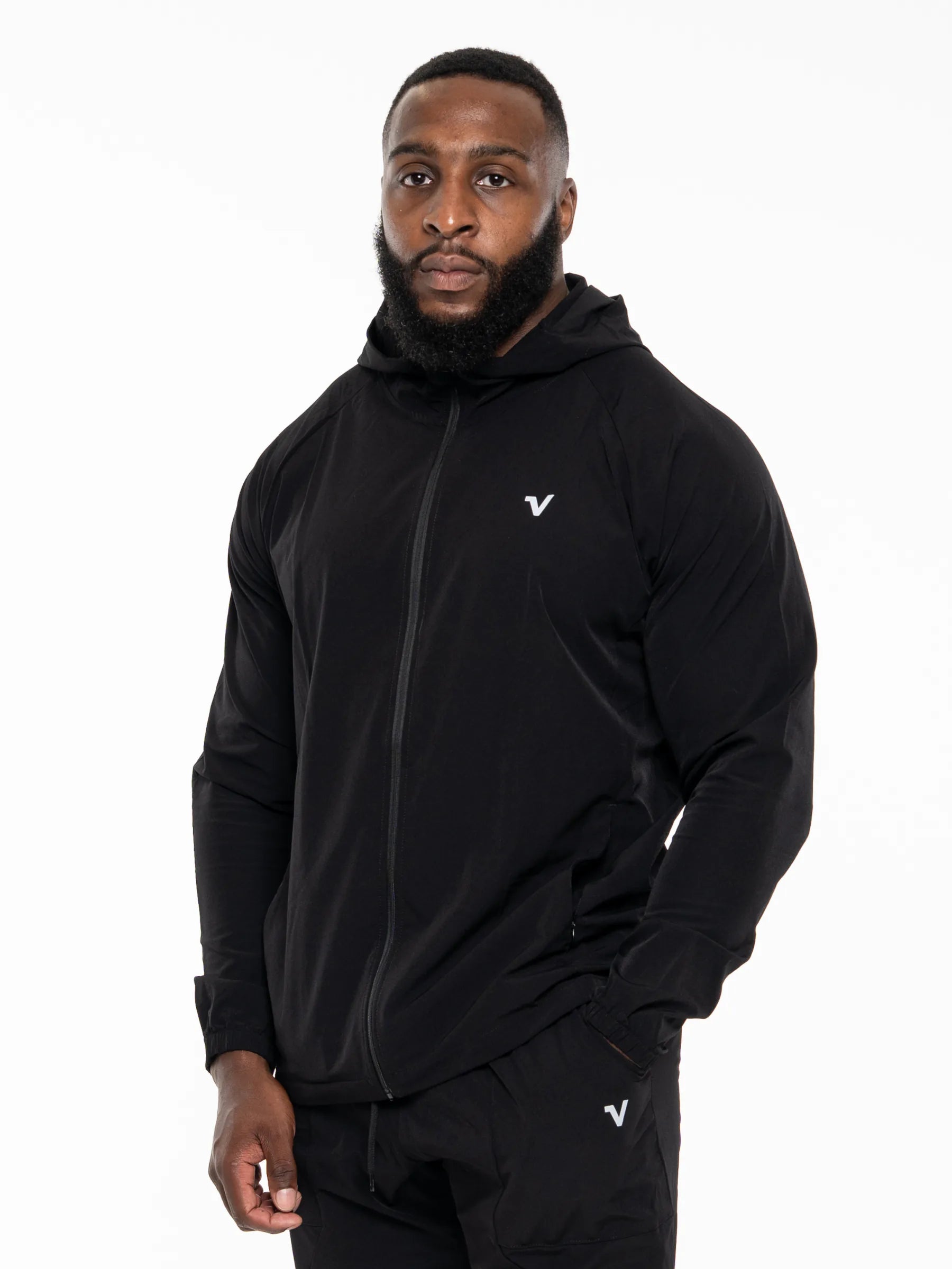 Veritone Mode Jacket, Midnight Black – A man wearing a black full-zip hooded jacket and matching black pants with a small white V logo on the chest and thigh, photographed against a plain white studio background.