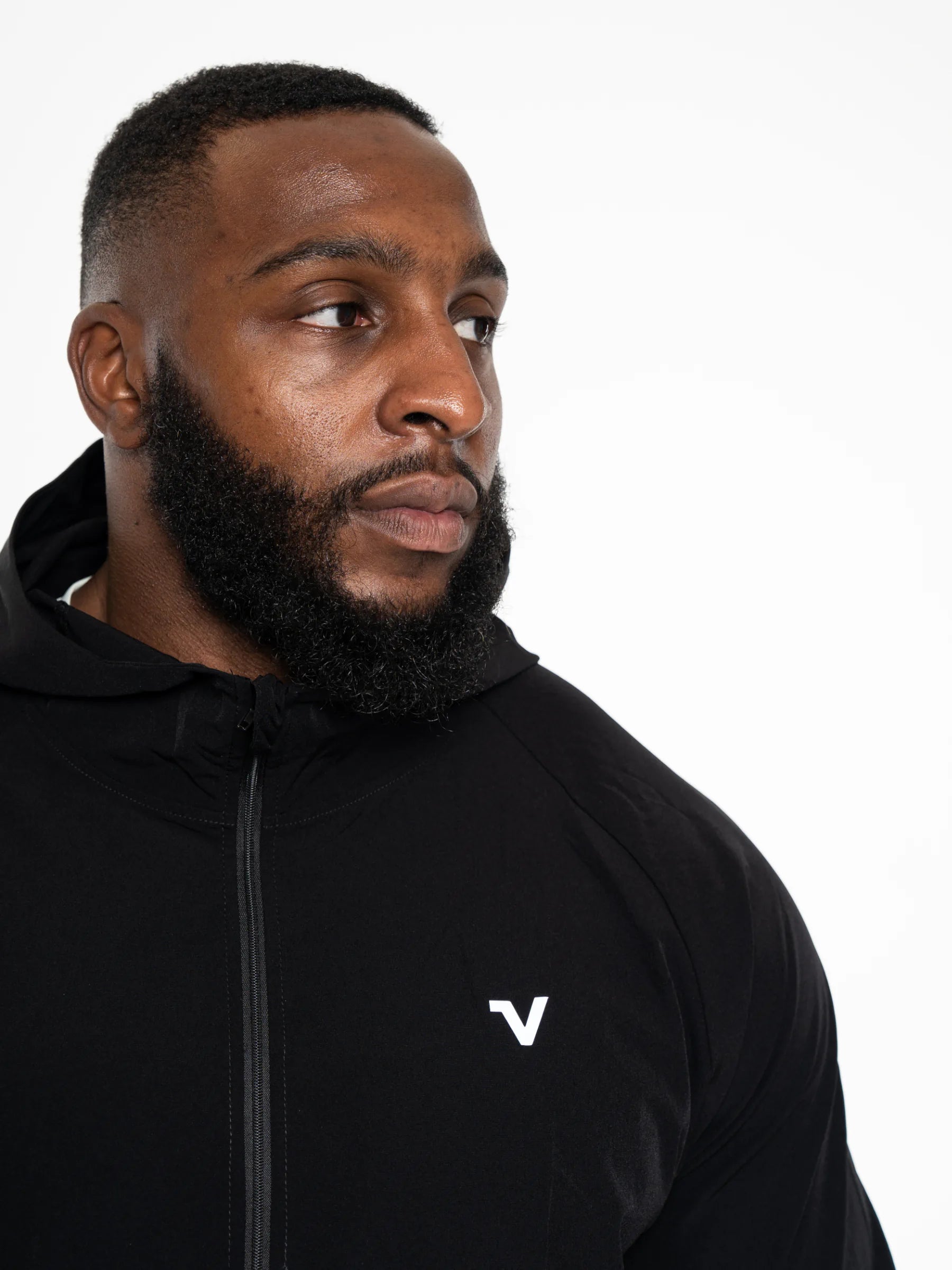 Veritone Mode Jacket, Midnight Black – A close-up of a man wearing a black athletic zip-up hoodie with a small white stylized 'V' logo on the left chest, photographed against a plain white studio background.