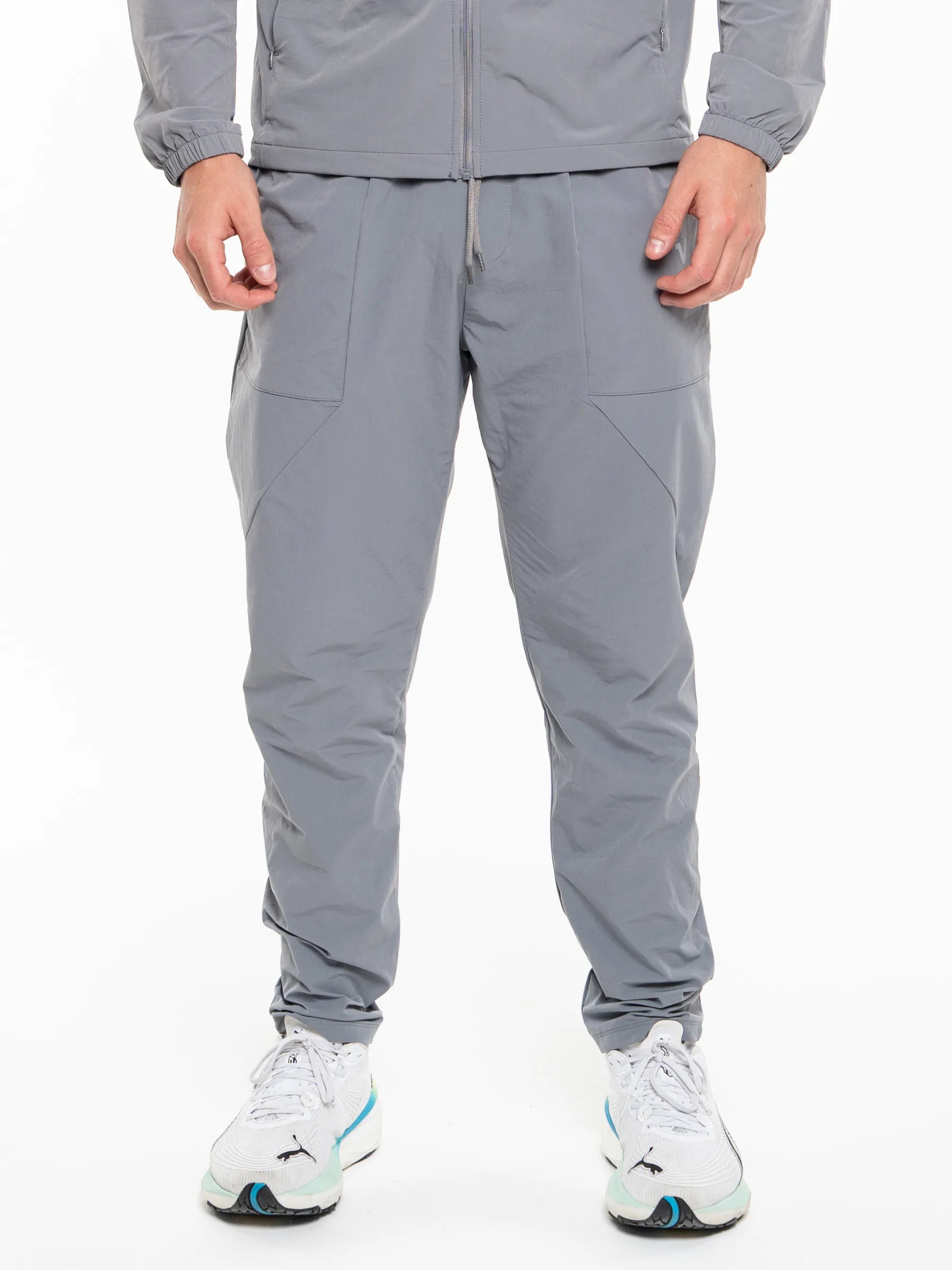 Veritone Mode Track Pants, Ash Grey – A person wearing light gray jogger-style athletic pants with a matching zip-up top and white sneakers, shown from the chest down against a plain white studio background.