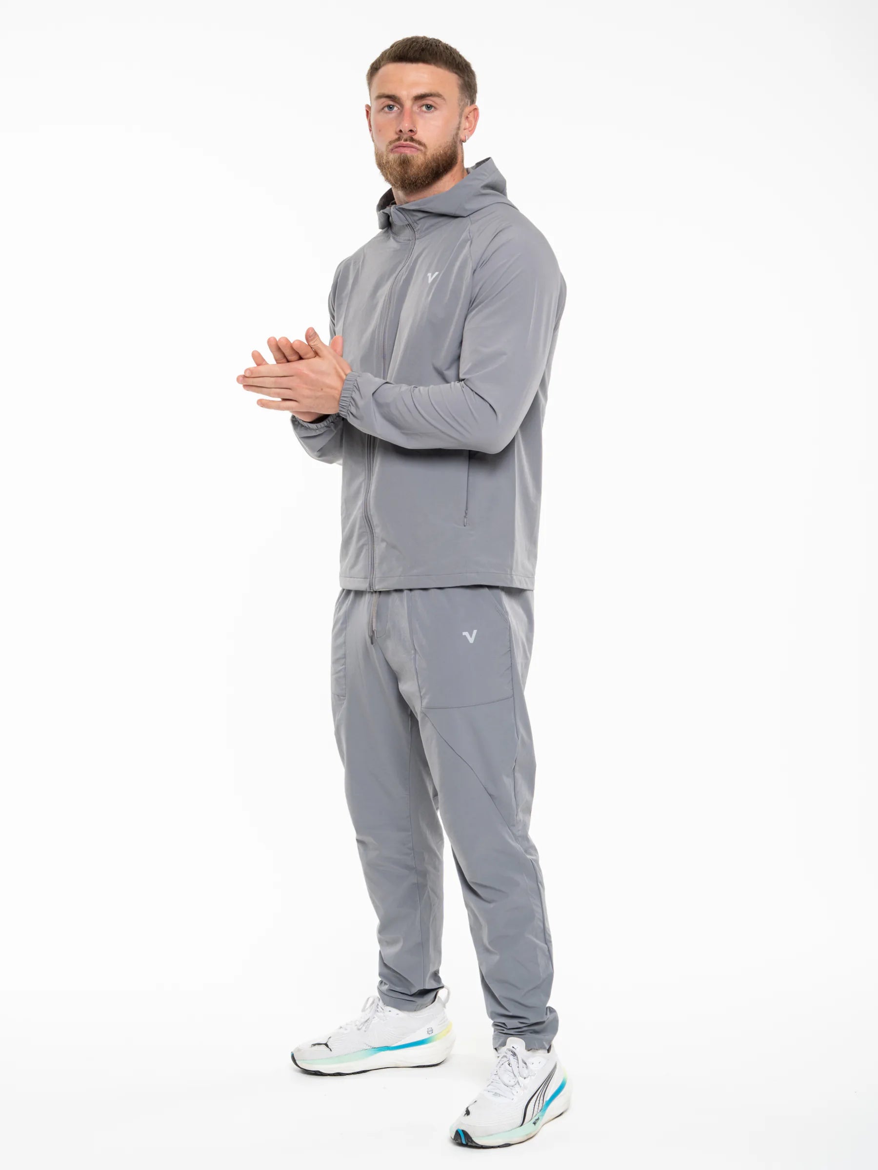 Veritone Mode Track Pants, Ash Grey – A man modeling a matching gray athletic tracksuit, featuring a zip-up hoodie and jogger pants, with small white embroidered logos on the chest and thigh, paired with white sneakers.