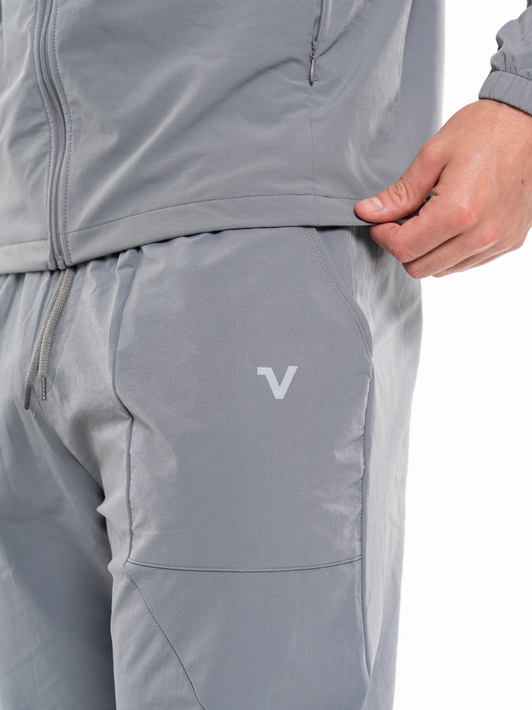 Veritone Mode Track Pants, Ash Grey – Close-up of a person wearing light gray athletic pants, pulling the side seam outward with one hand. A small white stylized 'V' logo is visible on the front pocket.