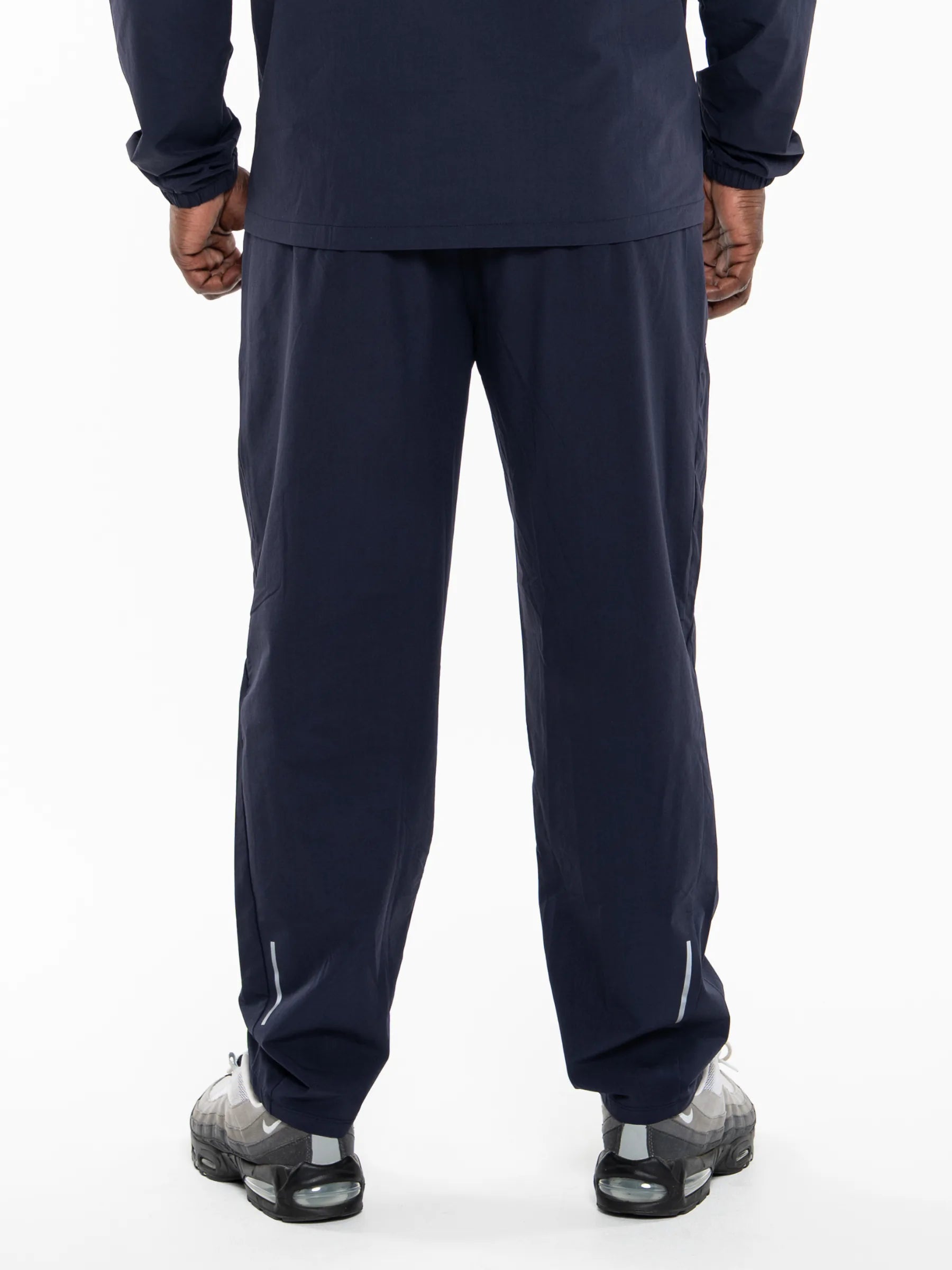 Veritone Mode Track Pants, Deep Navy – Rear view of a person wearing navy blue athletic pants and a matching navy long-sleeve top, paired with gray and black sneakers, against a plain white studio background.