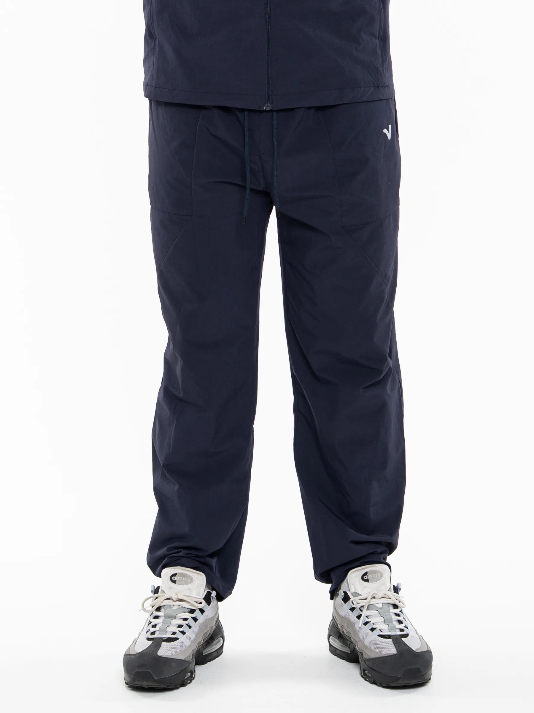 Veritone Mode Track Pants, Deep Navy – A person wearing dark navy pants with a small white logo on the left thigh, paired with gray and black sneakers, shown from the waist down against a plain white background.