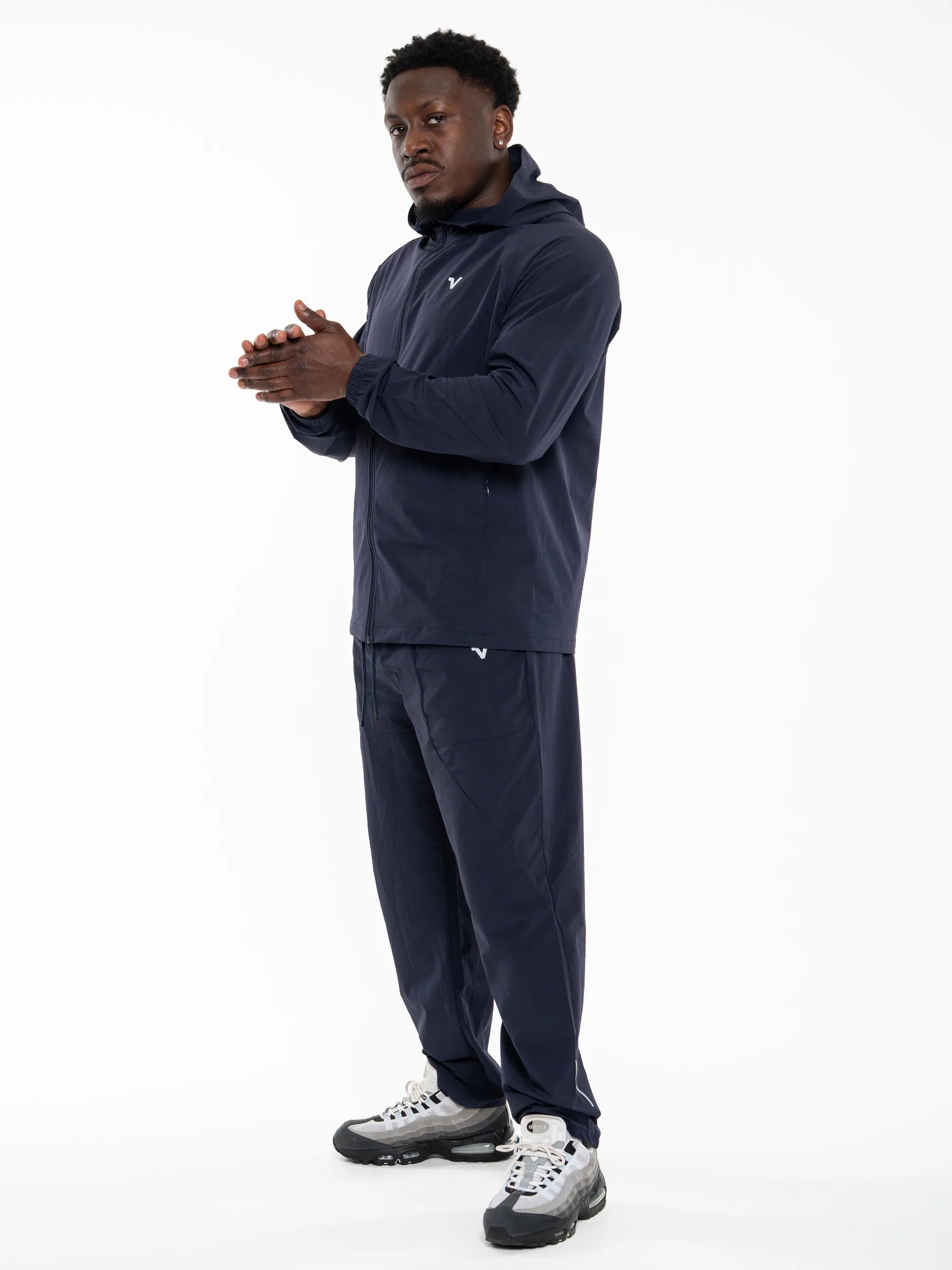 Veritone Mode Track Pants, Deep Navy – A man modeling a matching navy blue hooded sweatshirt and jogger pants set, paired with gray and black athletic sneakers, against a plain white studio background.