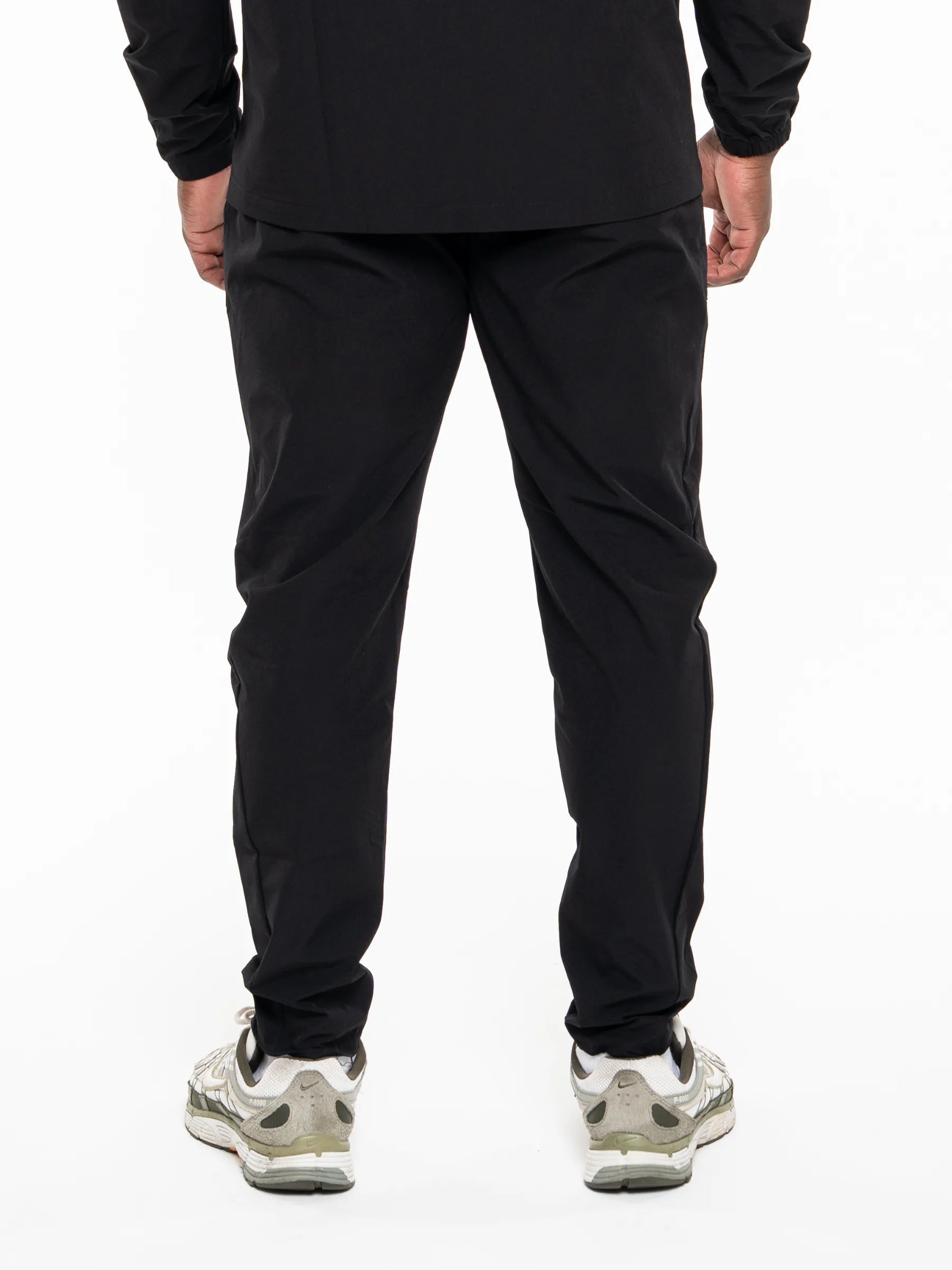 Veritone Mode Track Pants, Midnight Black – Rear view of a person wearing black athletic pants and white sneakers against a plain white studio background.