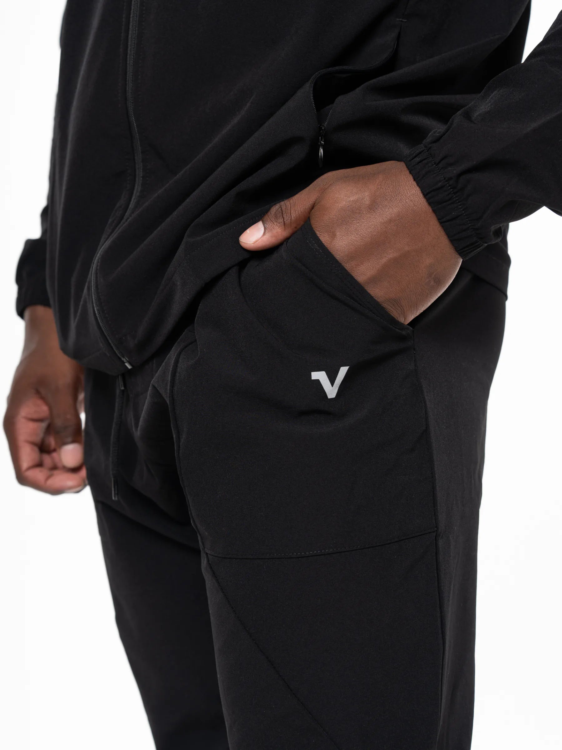 Veritone Mode Track Pants, Midnight Black – Close-up of a person wearing black athletic pants with a small white stylized V logo on the pocket, paired with a black zip-up top against a white studio background.