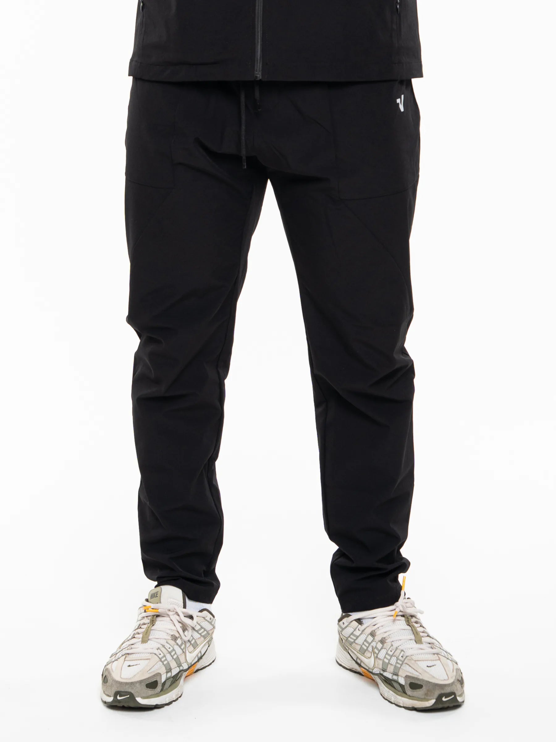 Veritone Mode Track Pants, Midnight Black – Black athletic jogger pants worn by a model, paired with white and gray sneakers, shown from the waist down on a white studio background.