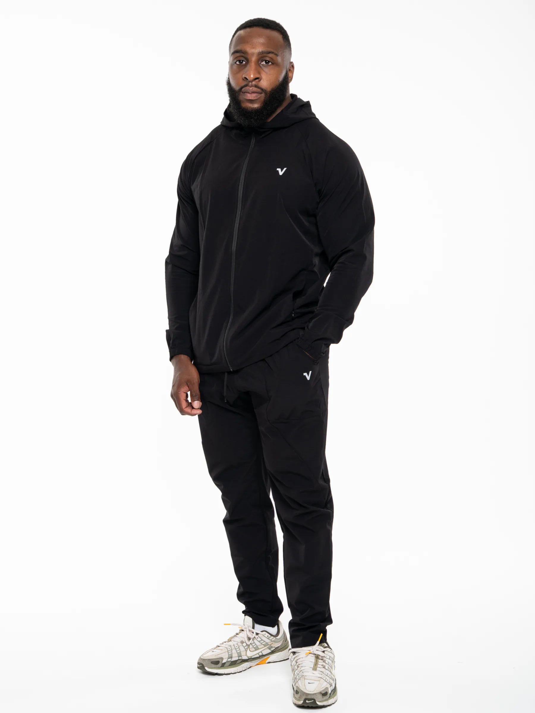 Veritone Mode Track Pants, Midnight Black – A man modeling a black zip-up hoodie and matching black jogger pants with small white logo accents, paired with gray and white sneakers, against a plain white studio background.