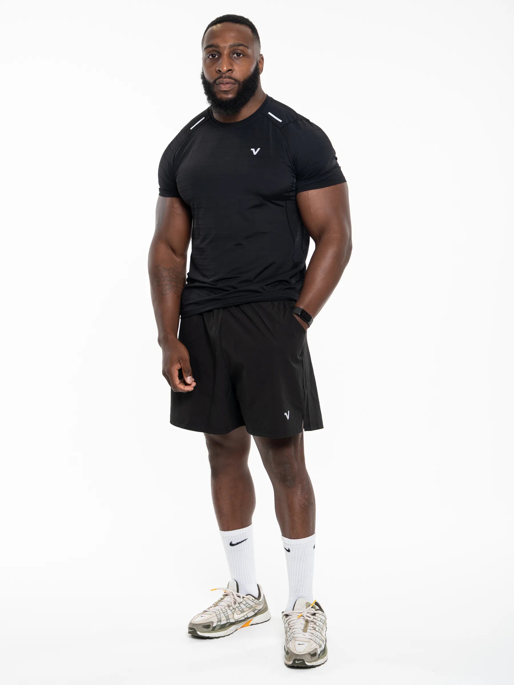 Veritone Prime Muscle Fit T-Shirt, Black - A muscular man modeling a black athletic t-shirt and matching black shorts, paired with white Nike socks and gray running shoes, against a solid white background.