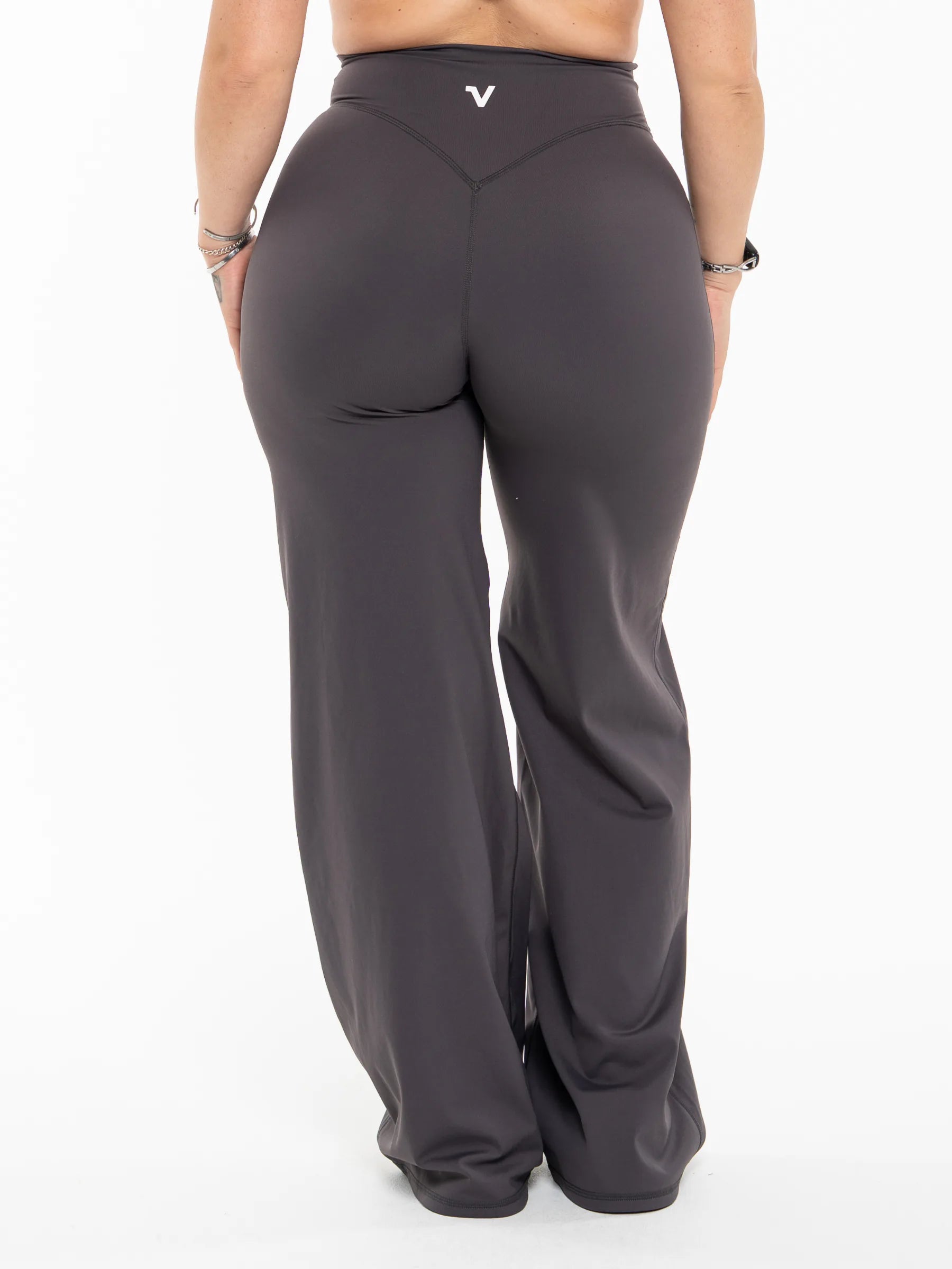 Veritone Sculpt Flared Pants, Charcoal Grey – A person shown from the back wearing dark charcoal gray wide-leg athletic pants with a small white logo on the waistband, photographed against a clean white studio background.